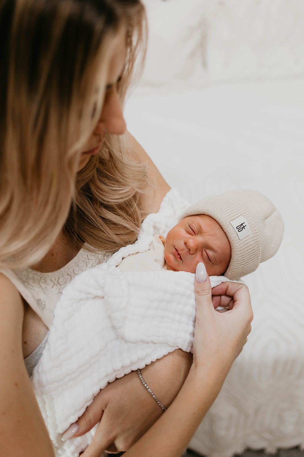 A woman with long, blonde hair tenderly holds a newborn baby wrapped in Forever French Baby's Crib Sheet in the Cloud pattern. The baby, wearing a beige knit hat, sleeps peacefully in her arms. The background is softly out of focus, highlighting the intimate and serene moment.