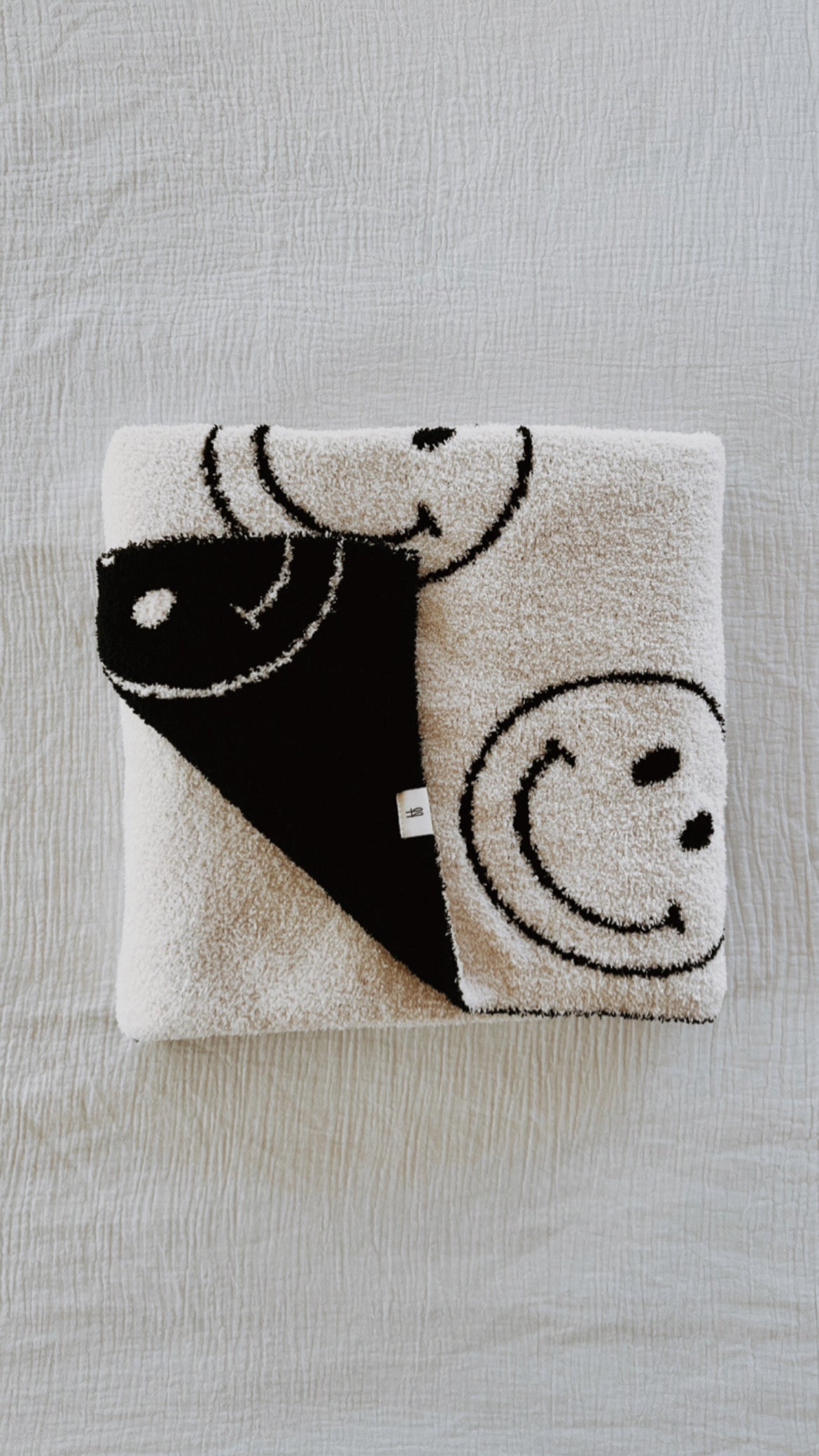 The Plush Blanket | Just Smile Black & White by forever french baby, featuring black smiley face designs on a white surface and a contrasting black corner with a white smiley face design, placed on a textured white background, evokes the cozy feel of a dreamy soft blanket.