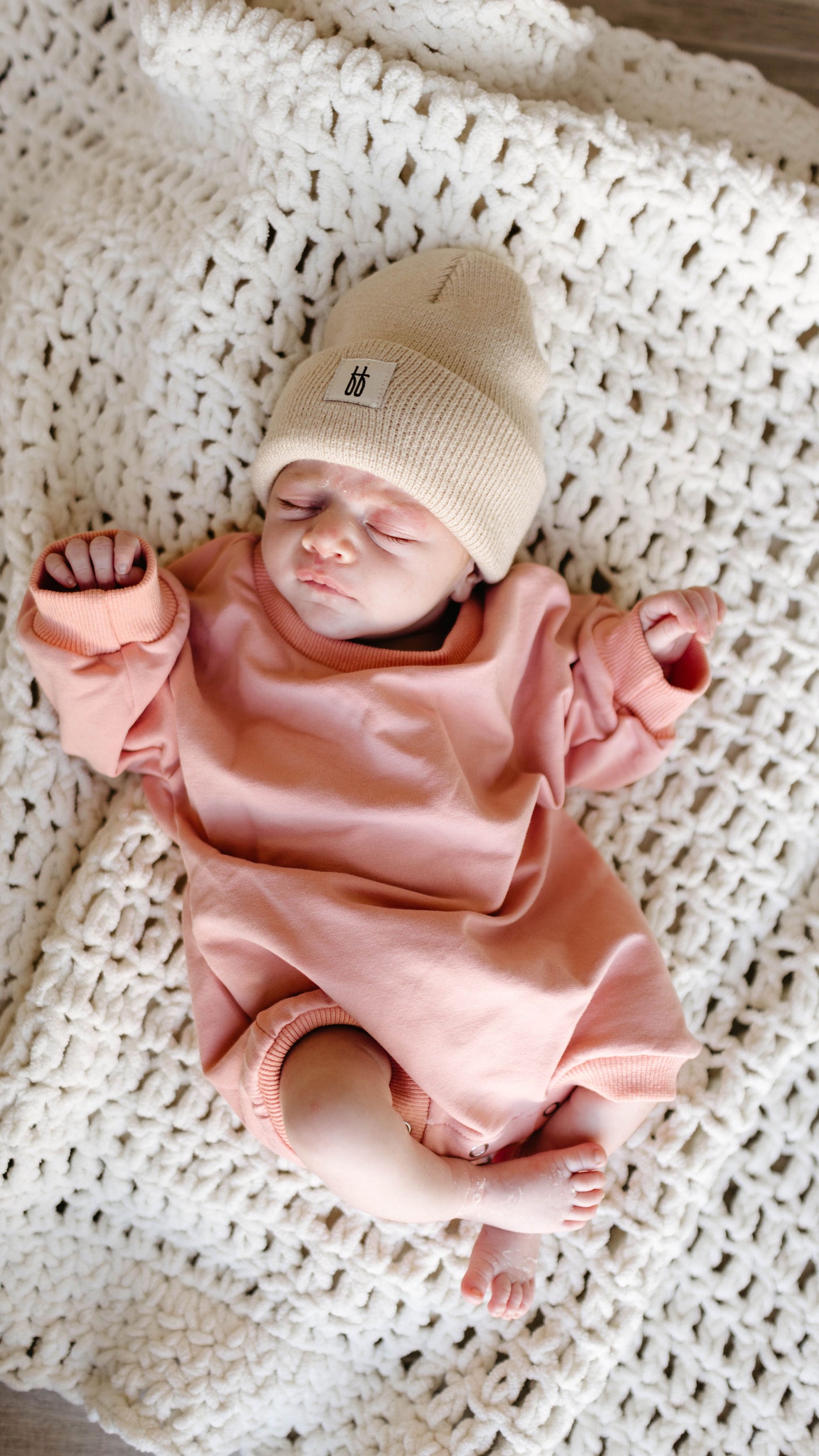 A newborn baby in the forever french baby Sweatshirt Romper | Rose rests on a textured white blanket, eyes closed and hands slightly raised, radiating pure innocence.