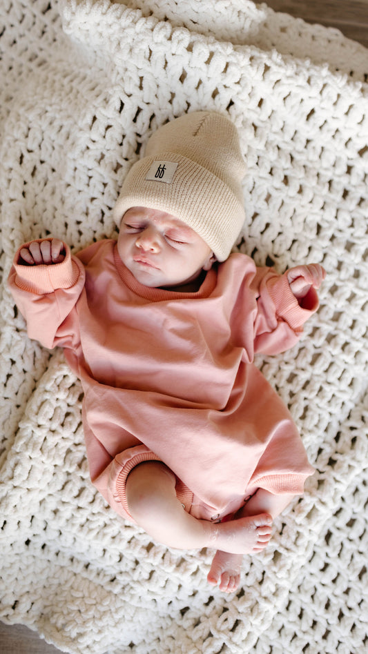 A newborn baby in the forever french baby Sweatshirt Romper | Rose rests on a textured white blanket, eyes closed and hands slightly raised, radiating pure innocence.