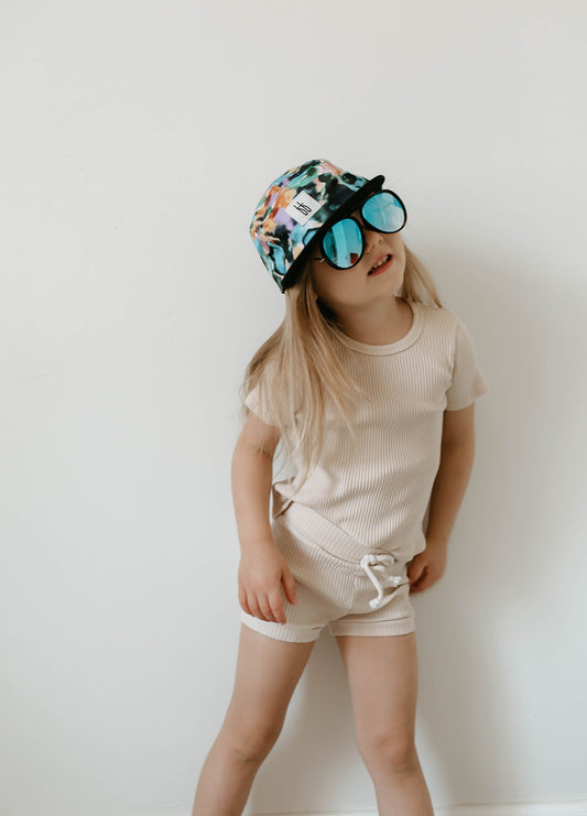 A young child stands against a plain white wall, wearing a beige outfit consisting of a short-sleeved shirt and shorts. The child sports the exclusive Snap Back | Charli Print cap from our limited collection by forever french baby and oversized mirrored sunglasses, looking stylish and cheerful with long hair flowing freely.