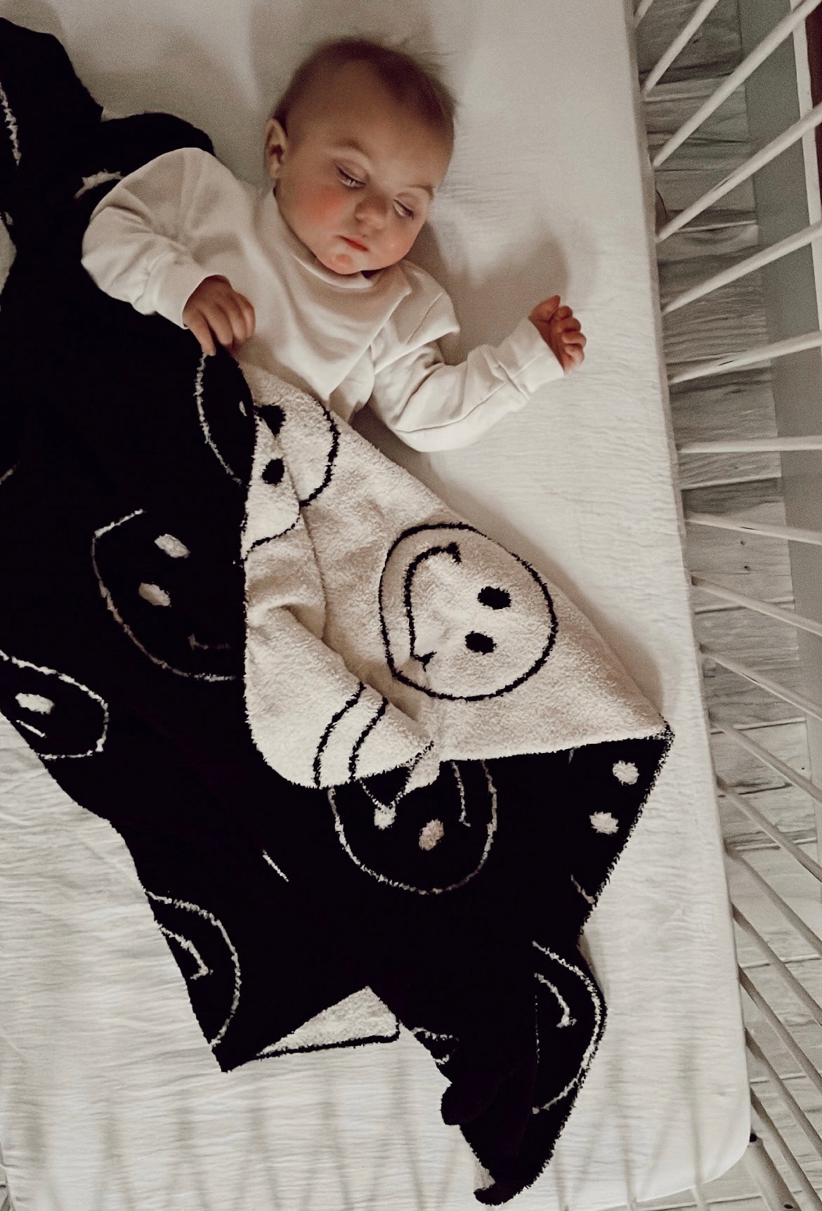 A baby peacefully sleeps in a crib, wrapped in the dreamy softness of the "Plush Blanket | Just Smile Black & White" from forever french baby, adorned with smiley face designs. The baby is dressed in a white long-sleeve outfit and lies on a white sheet. The crib bars are visible on the right side of the image.