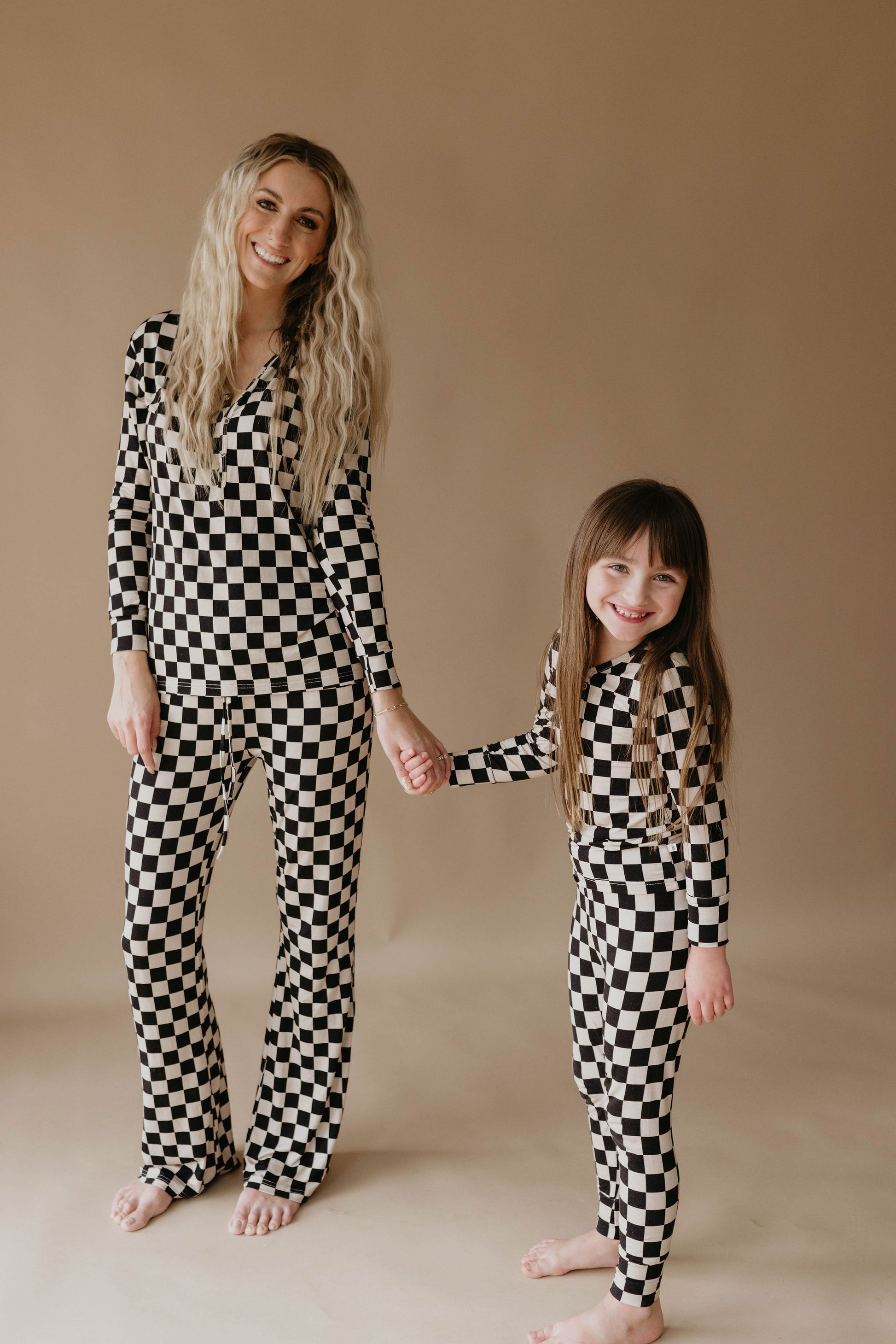 A woman and a young girl, both with long hair, stand holding hands against a plain background. They are both smiling and wearing matching checkered one-piece outfits from 'forever french baby' called Women's Bamboo Pajamas in Black Checkerboard.