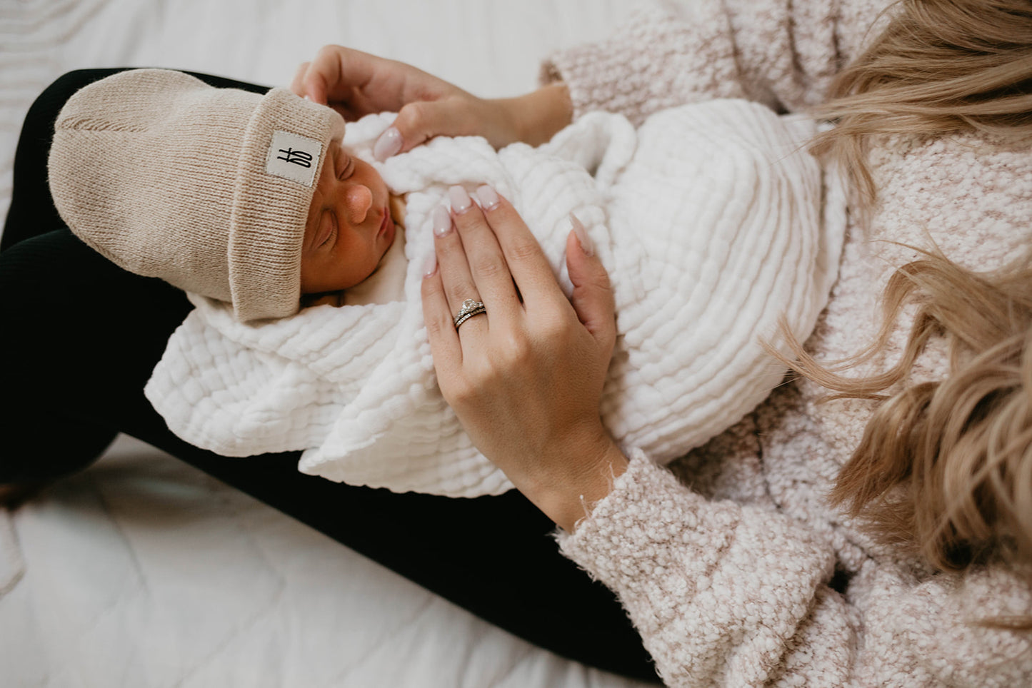 A close-up photo shows a person cradling a newborn baby wrapped in a cozy white blanket. The baby, wearing a beige knit hat from Forever French Baby and lying on the soft "Crib Sheet | Cloud" by Forever French Baby, is gently held. The person's hand, adorned with a ring and blond hair visible, adds to the tender moment—just like cherished garments washed with like colors.