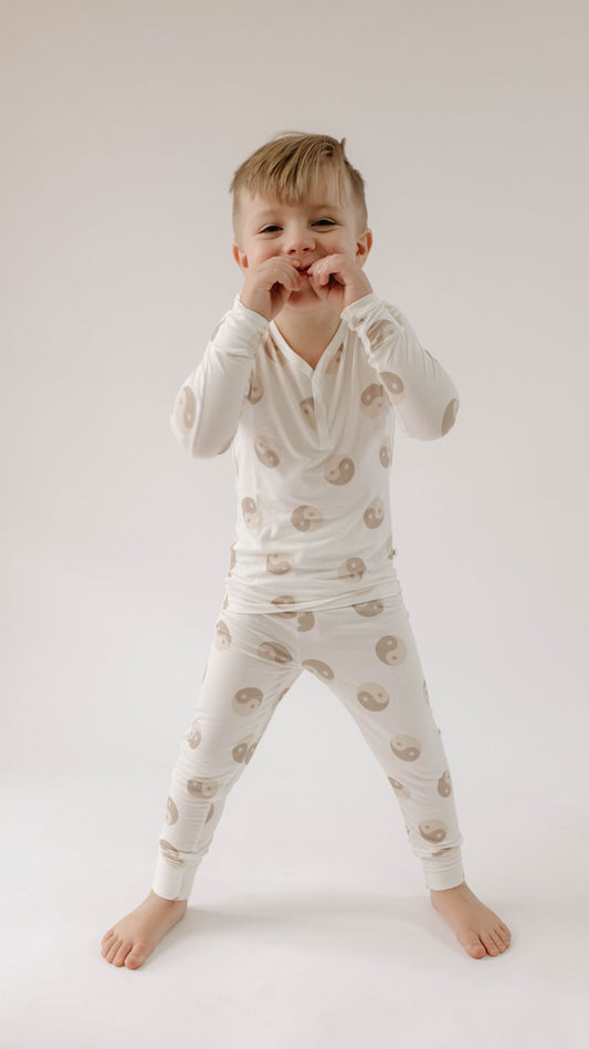 A young child stands and makes a playful hand gesture. The child is wearing forever french baby's hypo-allergenic Yin Yang Tan and White | Bamboo Two Piece Pajamas, adorned with a pattern of beige yin-yang symbols. The background is plain and white, giving the photo a clean and minimalistic look.