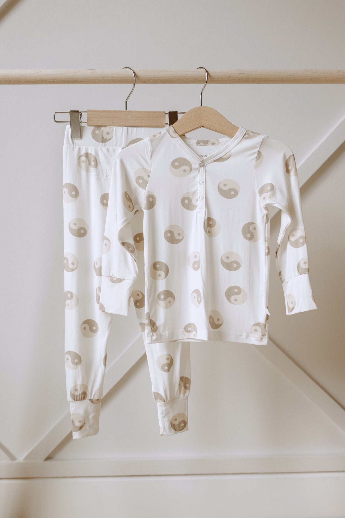 The Yin Yang Tan and White | Bamboo Two Piece Pajamas from forever french baby, a white long-sleeve set adorned with beige yin-yang symbols and crafted from hypo-allergenic bamboo fabric, is displayed on a wooden clothes rack against a light-colored, minimalist background. Perfect for your little one.