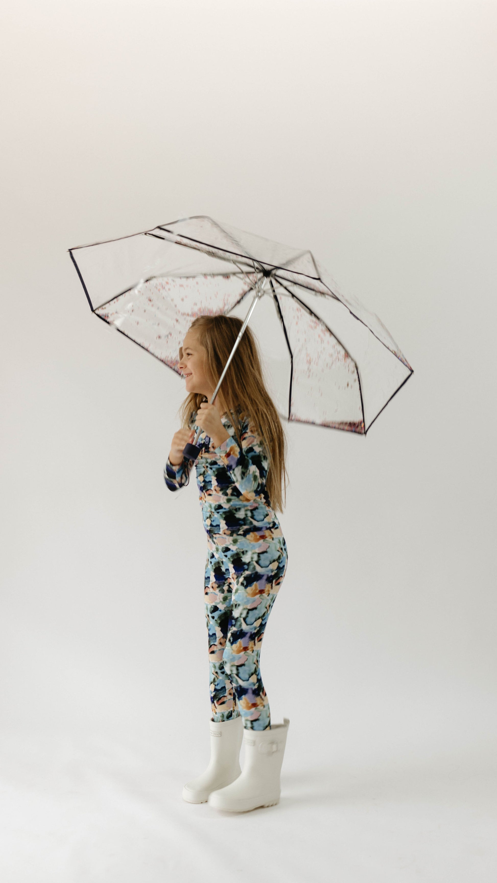 A young girl stands smiling under a transparent umbrella with red accents. She is wearing colorful, long-sleeved Bamboo Two Piece Pajamas in the Charli Print from forever french baby and white rain boots. The background is plain and white, giving a clean and simple look to the image.