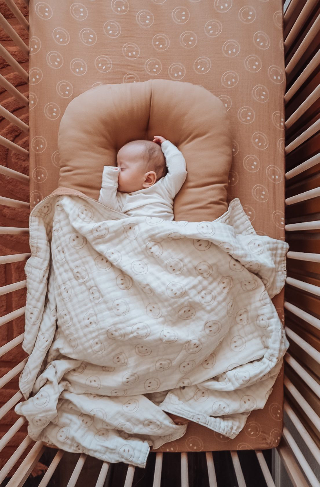 A baby sleeps peacefully in a wooden crib, wrapped in a Forever French Baby Muslin Quilt in Tan & Ivory Smile. The crib mattress is covered with an orange sheet featuring smiley faces. The baby rests under a tan, U-shaped pillow with one arm raised above the head.