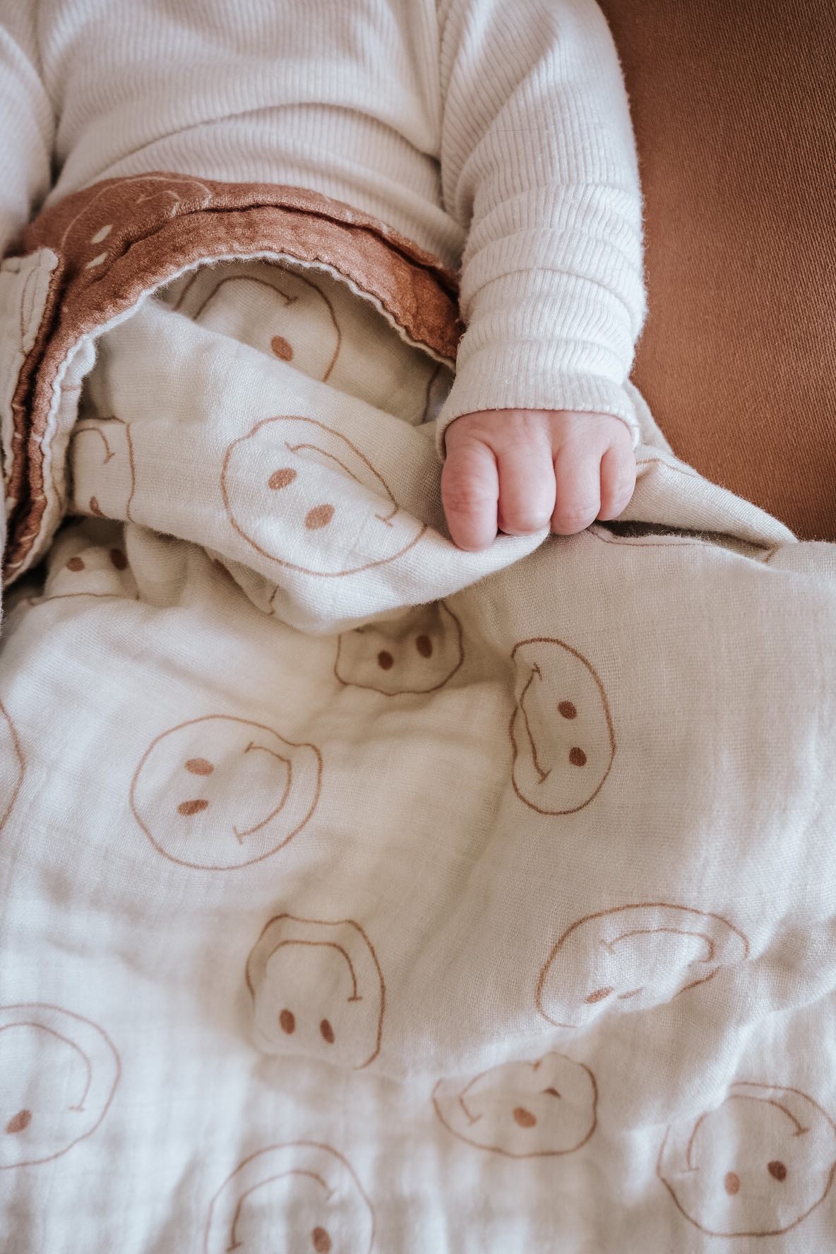 A baby wearing a white long-sleeve top is holding the "Muslin Quilt | Tan & Ivory Smile" from forever french baby. The quilt, adorned with brown smiley faces, is gently gripped in the baby's hand, creating a soft and cozy ambiance.