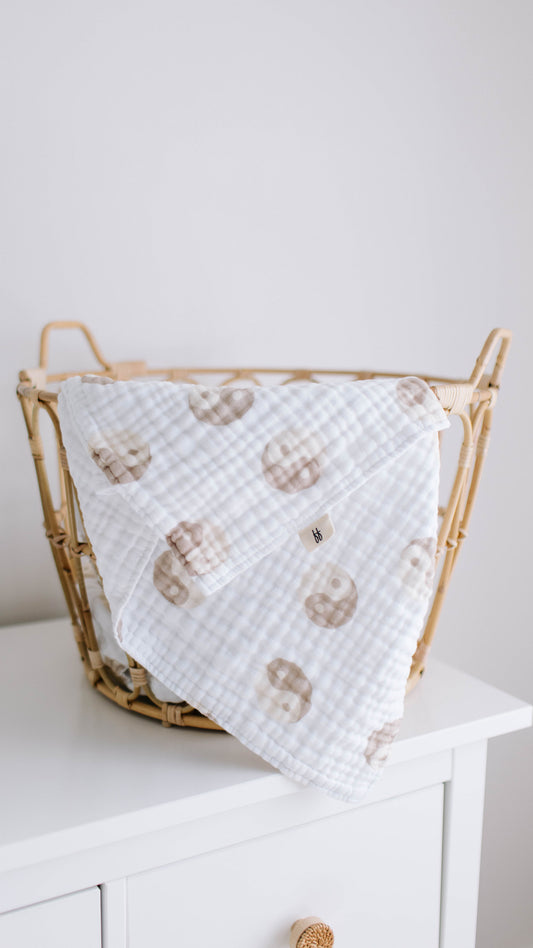 A wicker basket sits atop a white dresser, draped with the Yin Yang Quilt from Forever French Baby. The quilt is made from 100% muslin cotton and adorned with a cute, light brown sloth pattern—perfect for your little one. The background wall is plain and light-colored, creating a clean and minimalistic look.