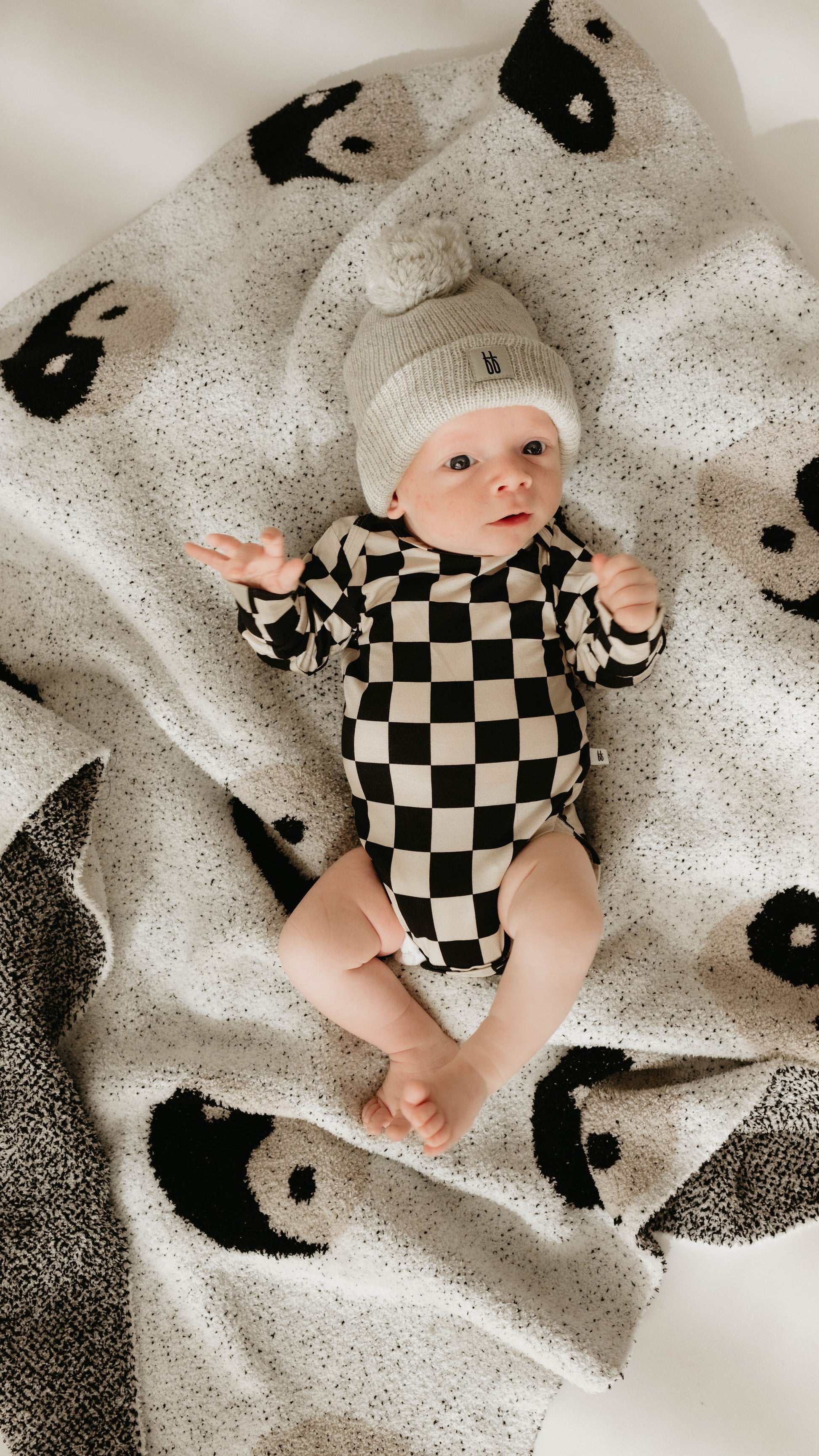 A baby wearing a black and white checkered onesie and a gray knit hat with a pom-pom lies on the forever french baby Plush Blanket in the Black & White Yin Yang design. The velvety-rich textured microfiber feather yarn of the blanket adds extra coziness as the baby looks up with wide eyes and has hands slightly raised.