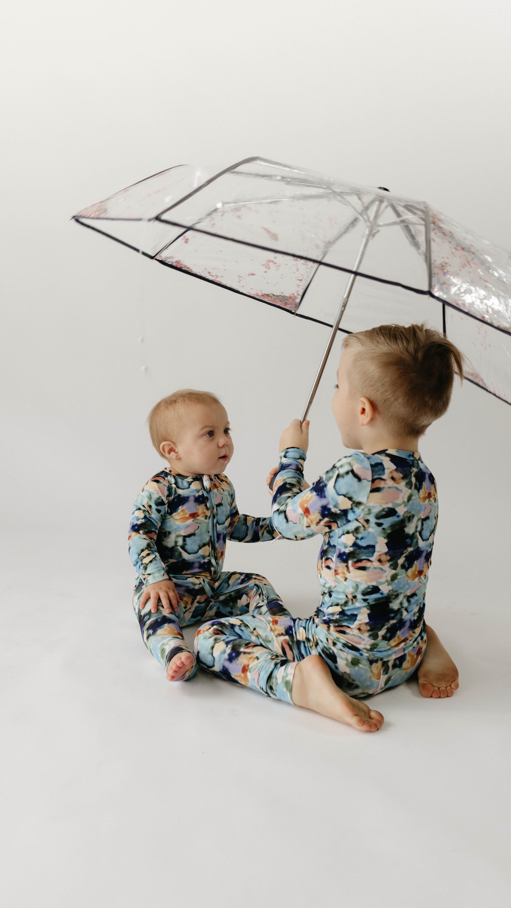 Two young children, both dressed in matching Charli Print Bamboo Two Piece Pajamas from forever french baby, sit on a white surface. The older child holds a transparent umbrella adorned with colorful prints over them. The younger child gazes at the older one with curiosity. The background is plain and white.