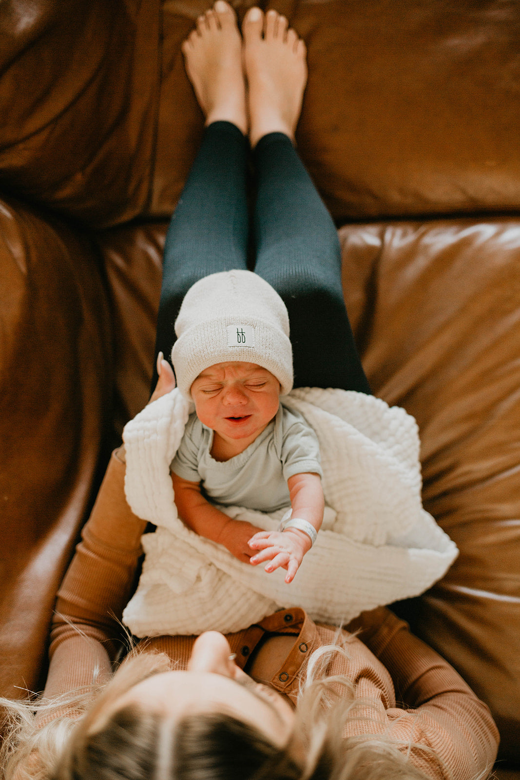 A person is seen from above lying on a couch with their legs extended. They are holding a crying baby wrapped in a white Forever French Baby Crib Sheet | Cloud. The baby is wearing a knit beanie and a light-colored onesie. The person’s face is partially visible, looking down at the baby.