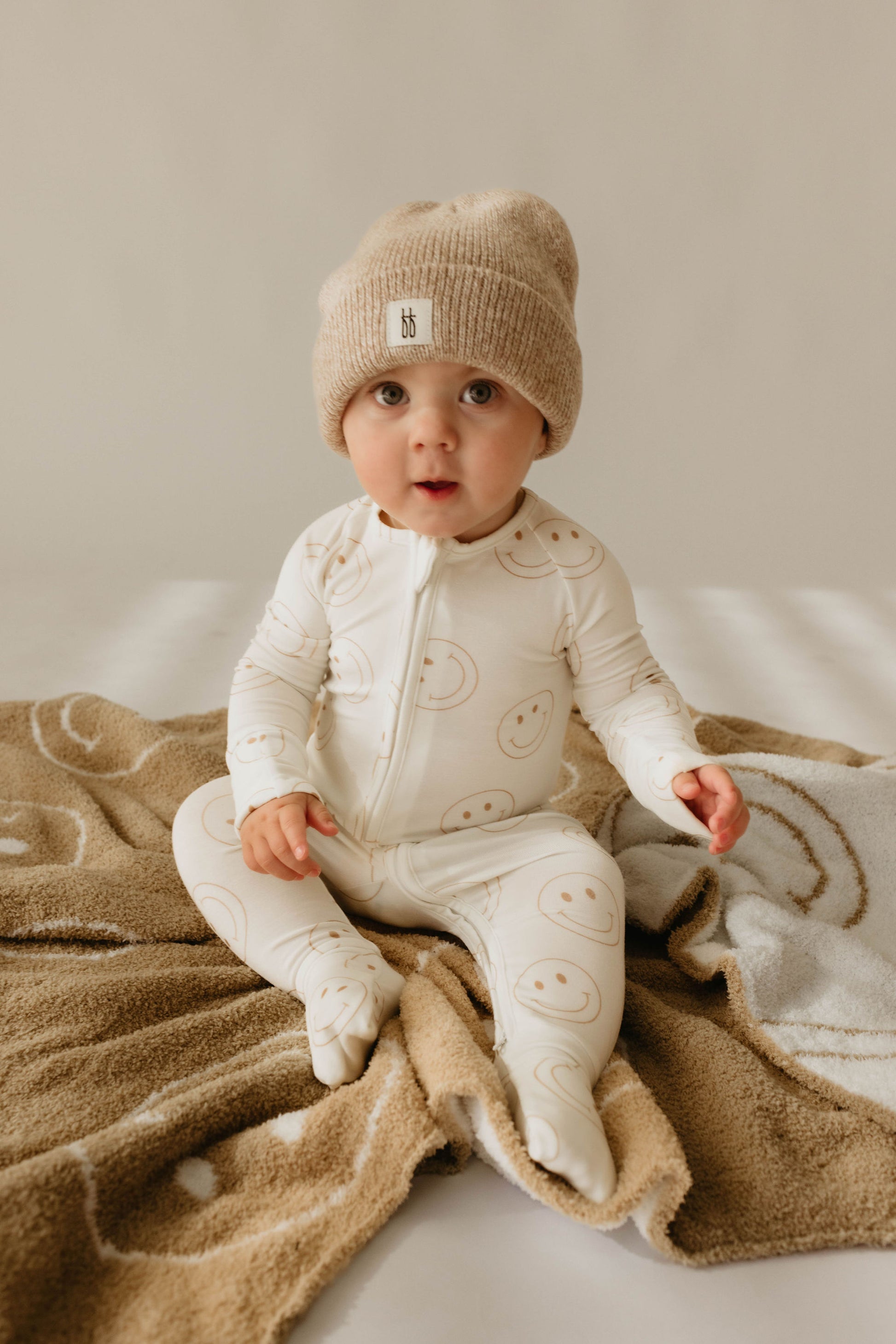 A baby in a beige knit beanie and white onesie with smiley faces from forever french baby sits on the Plush Blanket | Original Just Smile by forever french baby, looking at the camera against a neutral background.