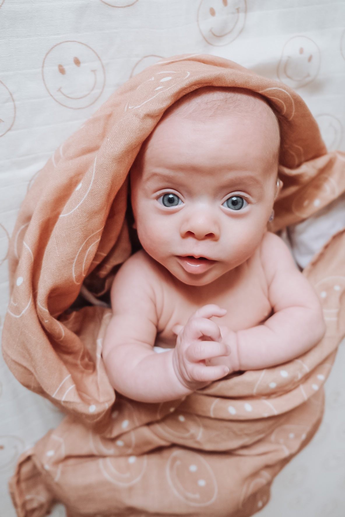 A baby with wide blue eyes is wrapped in the "just smile tan" muslin swaddle by forever french baby. The light brown swaddle features white smiley face patterns, and the child looks up with a curious expression, raising one hand near its face. The background is a white sheet adorned with matching smiley face patterns.