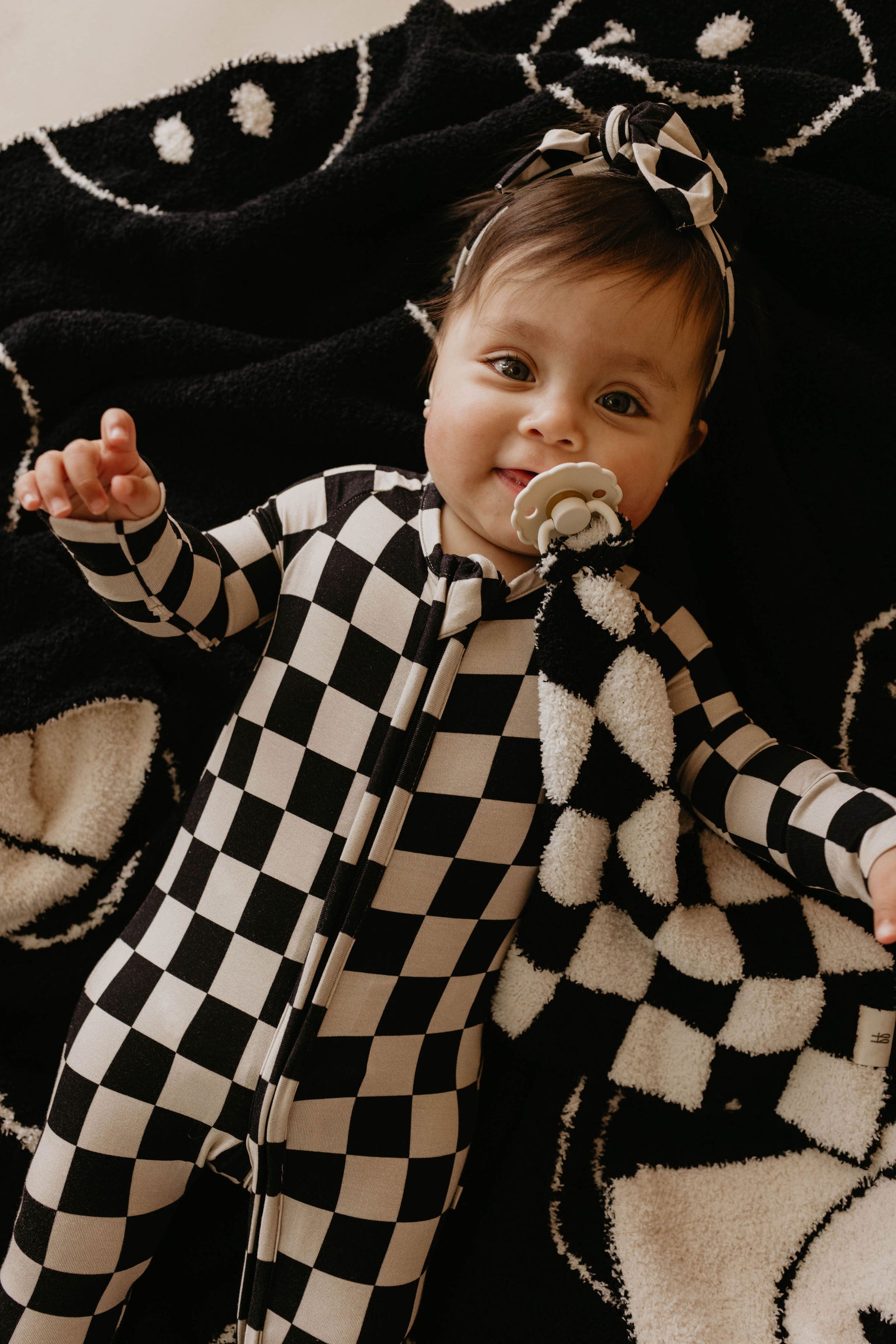 A baby with dark hair lies on a black and white patterned blanket. The baby is wearing a black and white checkerboard onesie and matching headband, holding a pacifier while clutching the Plush Blanket | Just Smile Black & White by forever french baby. The baby is smiling and looking up.