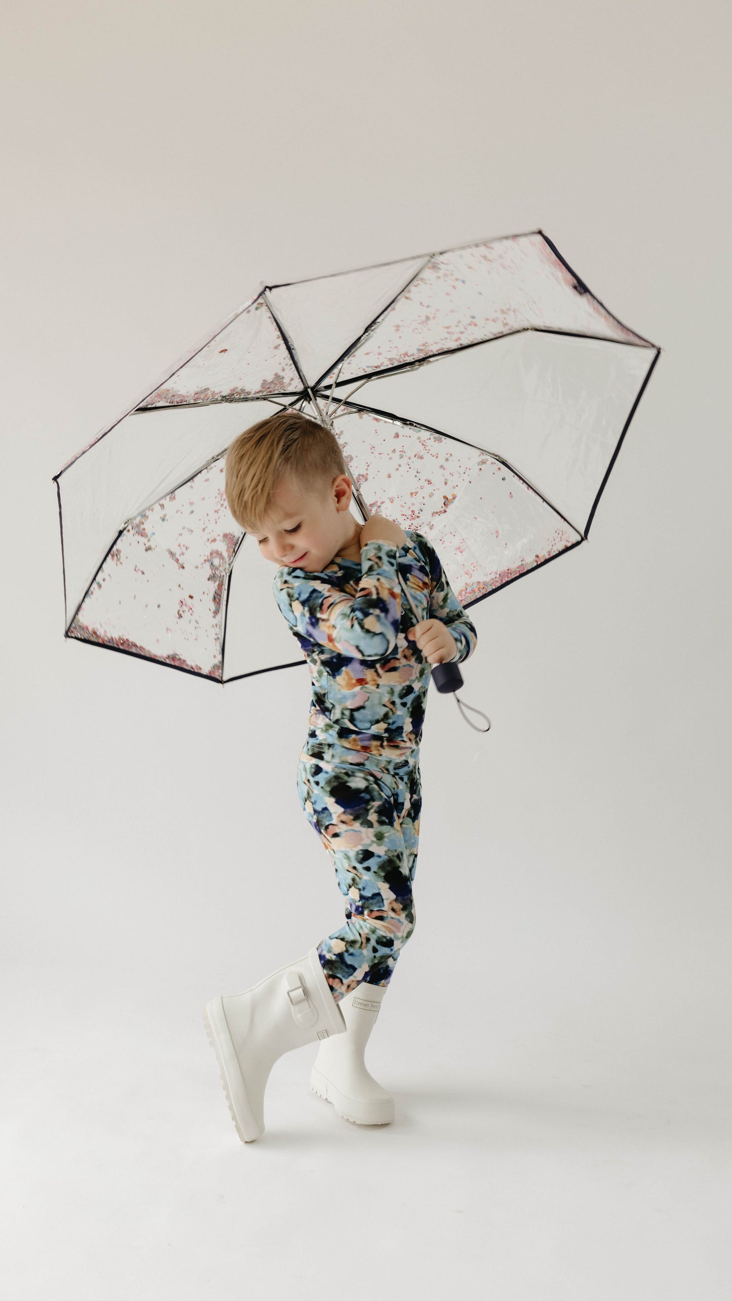 A young child stands on one foot, balancing a transparent umbrella overhead. The child is dressed in a colorful floral-patterned outfit called the Bamboo Two Piece Pajamas in Charli Print by forever french baby, paired with white boots. The background is plain and light-colored, making the child the focal point.