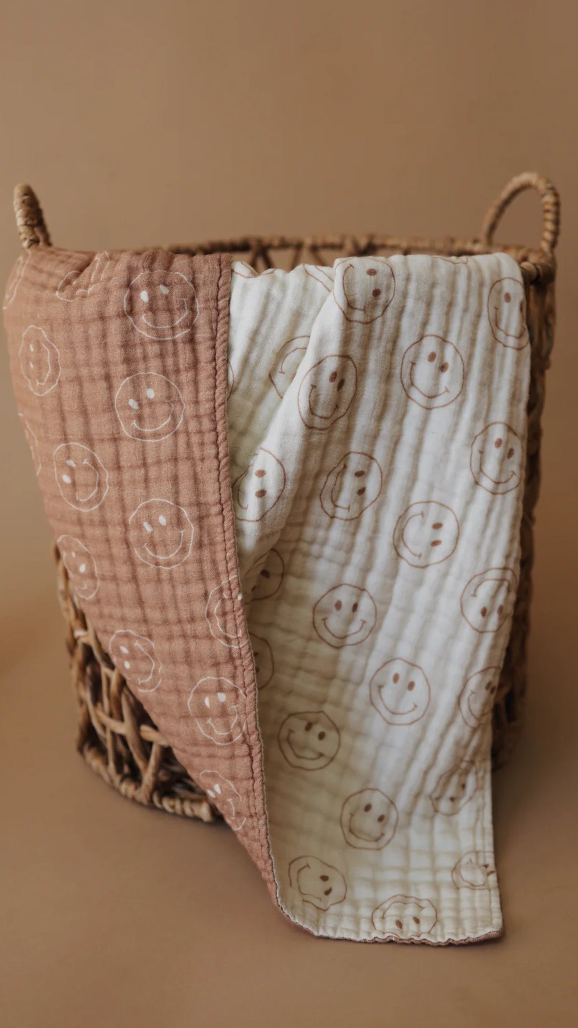 A woven basket rests against a neutral background. Draped over the basket is a Muslin Quilt | Tan & Ivory Smile from forever french baby, featuring two distinct sections: a light brown side and a white side, both adorned with smiling face patterns. The muslin cotton quilt appears soft and cozy.