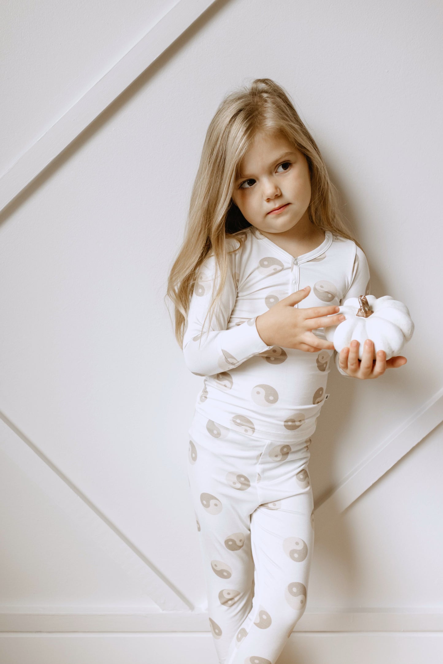 A young child with long blonde hair, dressed in the hypo-allergenic "Yin Yang Tan and White | Bamboo Two Piece Pajamas" by forever french baby, stands against a white wall. The child holds a small white pumpkin in their hands and gazes thoughtfully to the side.