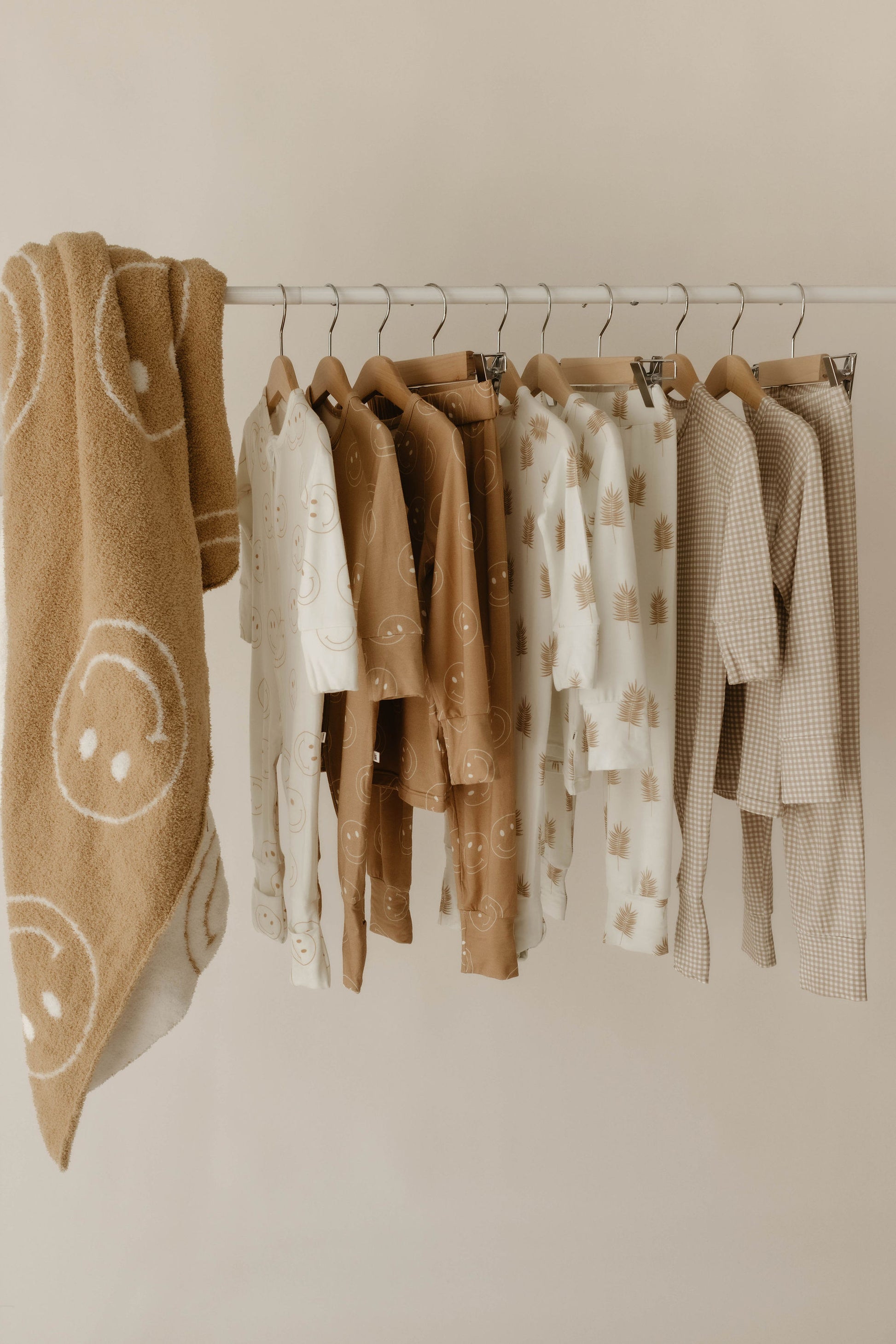 A clothing rack holds neutral-toned children’s clothes with smiley faces, palm trees, and checks next to the forever french baby Plush Blanket | Original Just Smile—dreamy soft with big white smiley faces—set against a plain light beige wall.