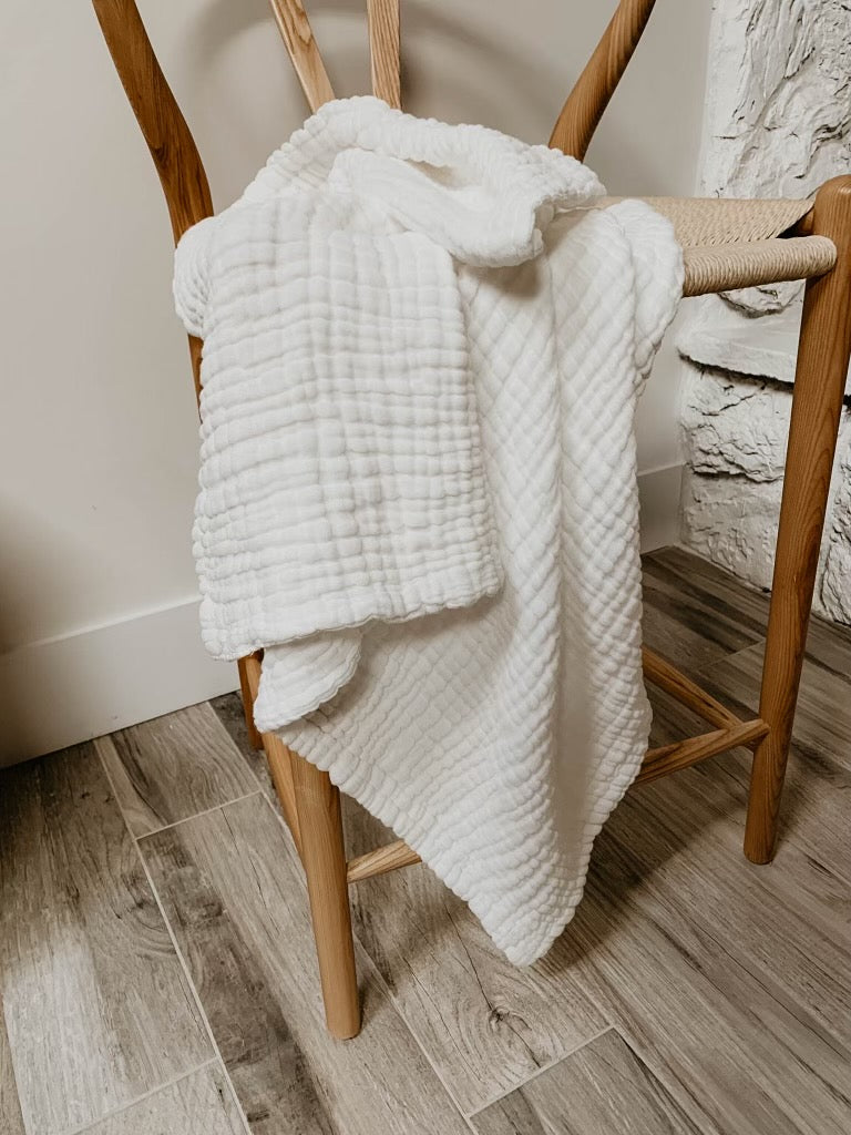 A cozy Crib Sheet | Cloud from forever french baby is draped over the back and seat of a wooden chair. The chair rests on a wooden floor, adjacent to a white stone wall in a snug indoor setting. For care instructions, wash with similar colors and tumble dry on low.