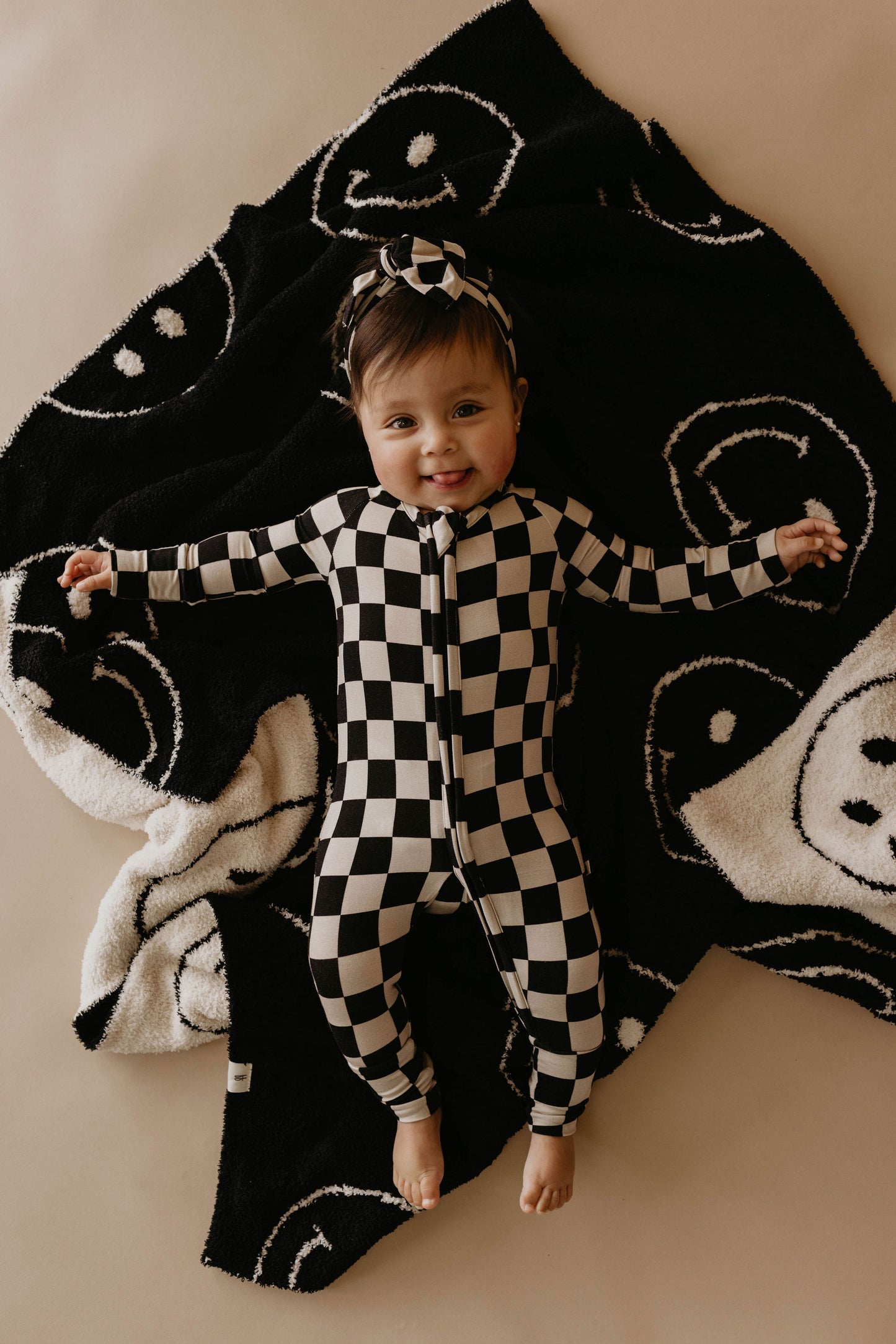 A smiling baby lies on the forever french baby's dreamy *Plush Blanket | Just Smile Black & White*, adorned with checkered patterns and smiley faces. The baby is dressed in a matching checkered onesie and headband, spreading their arms and legs out in a playful manner. The background is a light, neutral color.