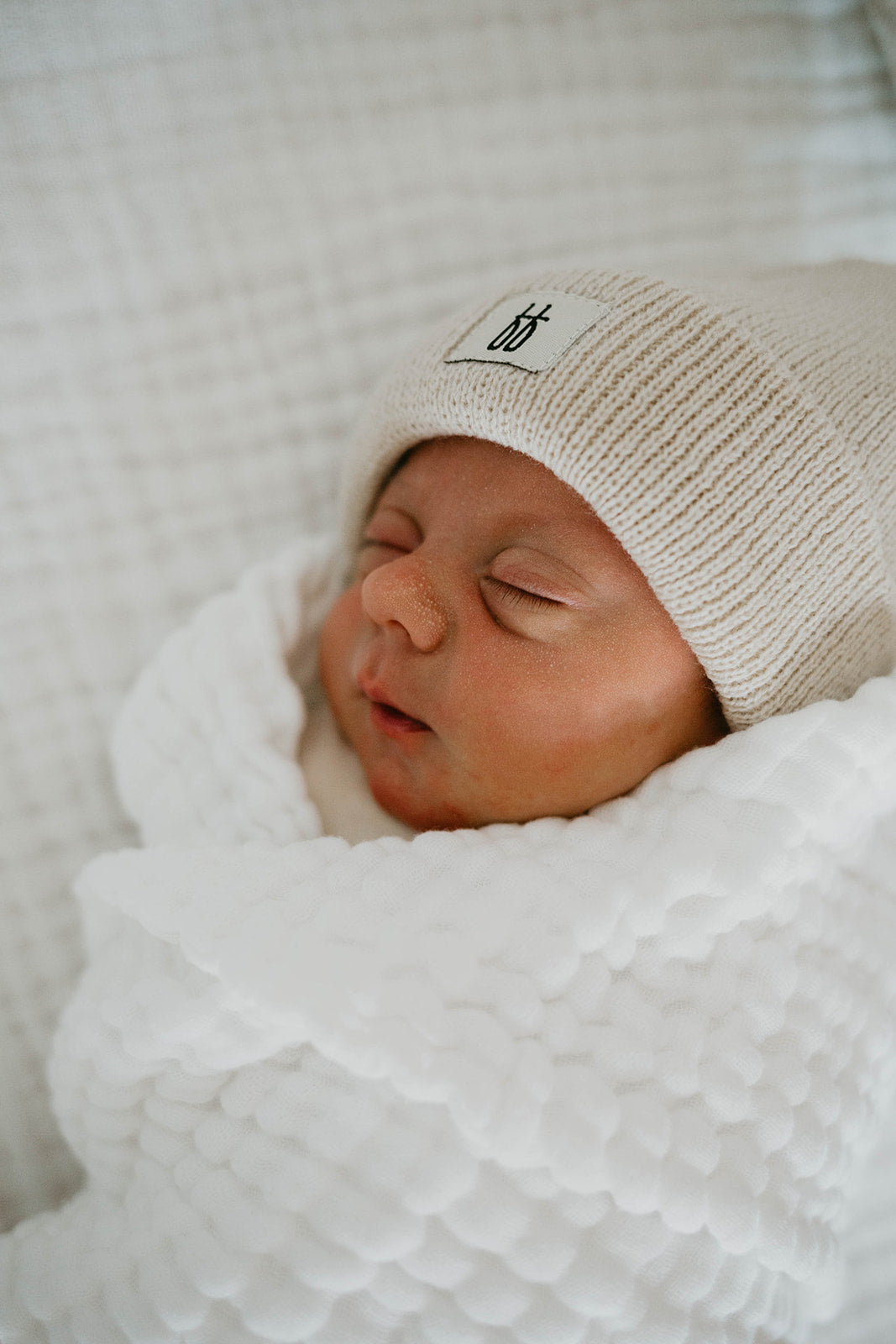 A newborn baby sleeps peacefully, wrapped in a cozy white blanket. The baby wears a light beige knit hat with a small forever french baby emblem on the front. The background features the soft and textured Crib Sheet in Cloud from forever french baby, complementing the gentle and serene atmosphere of the scene.