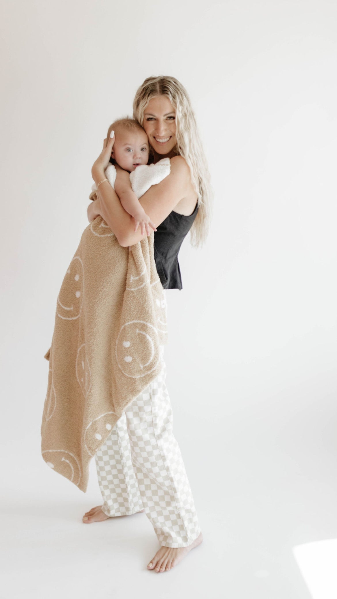 A person with long, wavy blonde hair smiles while holding a baby wrapped in the Plush Blanket | Just Smile Black & White by forever french baby. The person is wearing a black top and white checkered pants, standing barefoot against a plain white background.