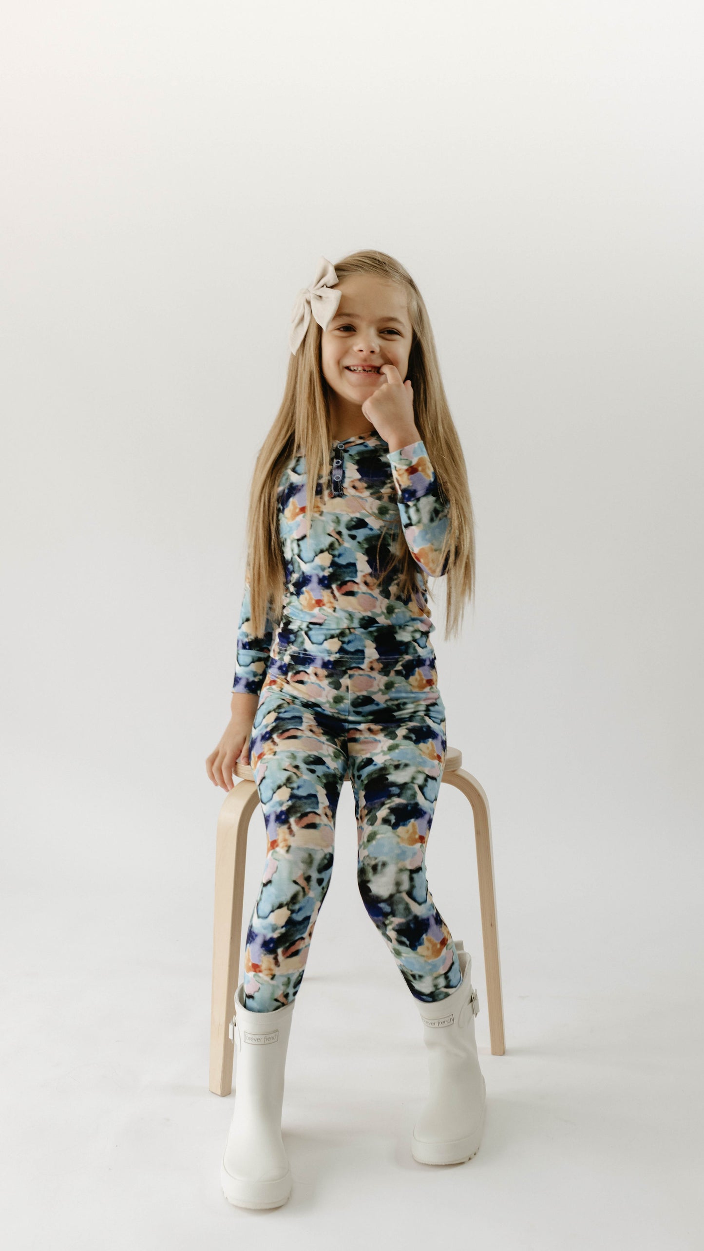 A young girl sits on a wooden stool against a plain, light background. She has long hair adorned with a large bow and is wearing forever french baby's Charli Print Bamboo Two Piece Pajamas, which consists of a long-sleeve top and leggings in a matching floral pattern. She is smiling with her hand touching her chin and wearing white boots.