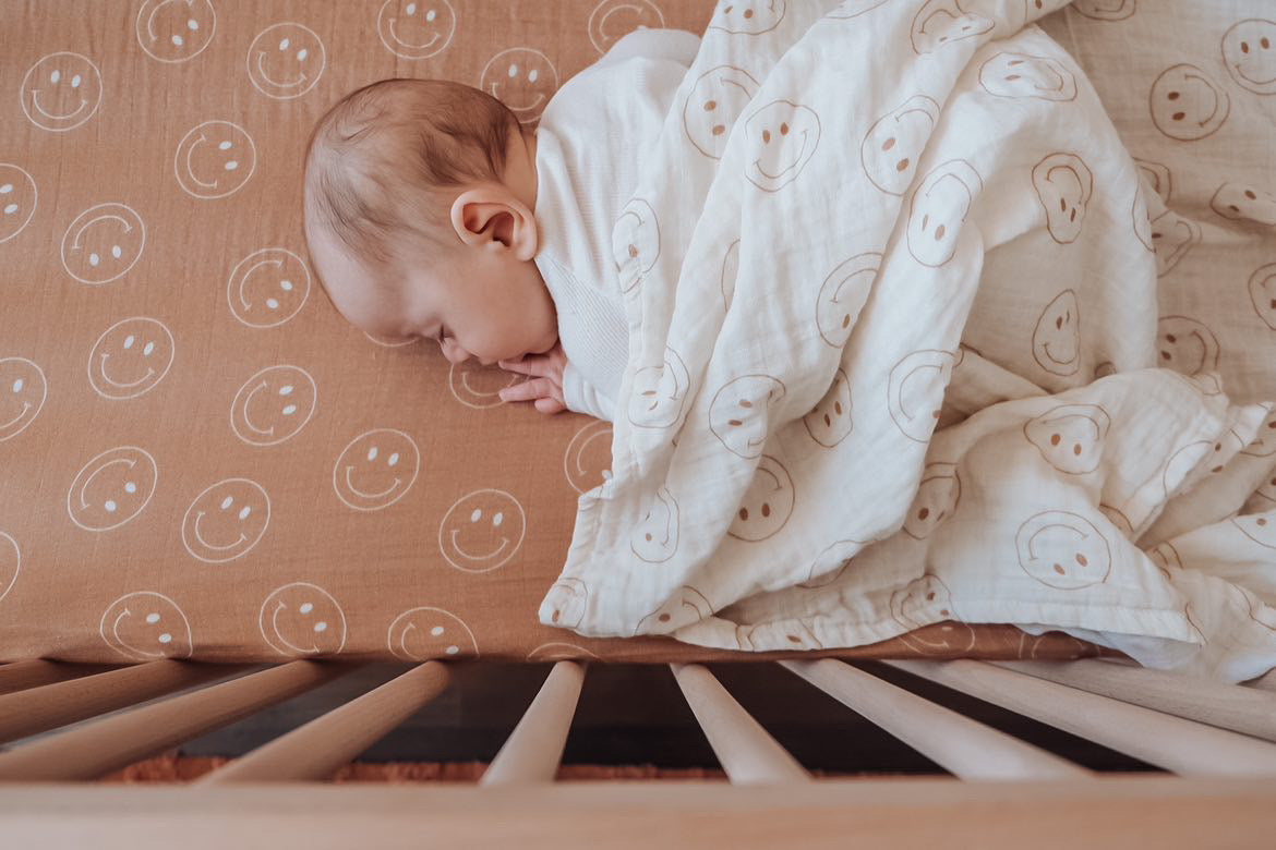 A baby sleeps soundly on a light brown sheet with smiley faces, covered by a Just Smile Ivory muslin swaddle from Forever French Baby that also features smiley faces. The crib's wooden slats are visible in the foreground, and the blanket is made of soft 100% muslin cotton.