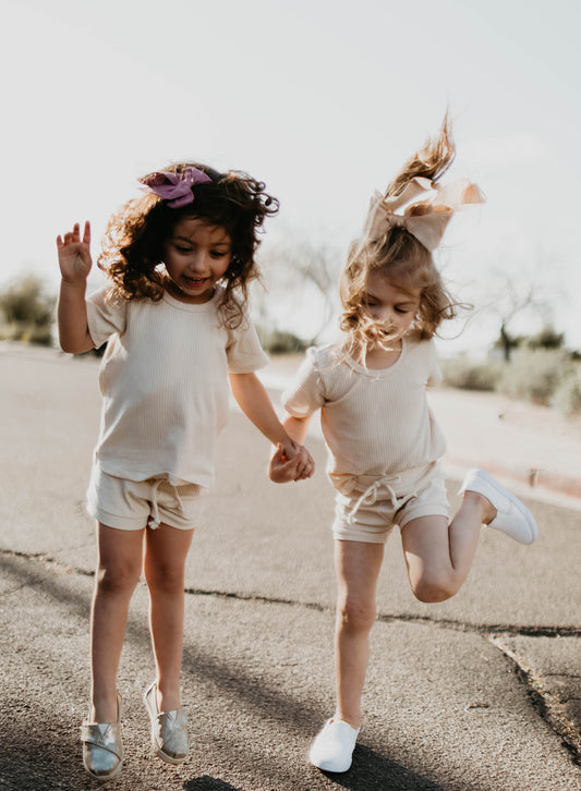 Two young girls joyfully hold hands while jumping on a sunlit pavement, both wearing forever french baby's Ribbed Set - Vanilla. One has dark curly hair with a purple bow, and the other has light hair in a ponytail with a bow.