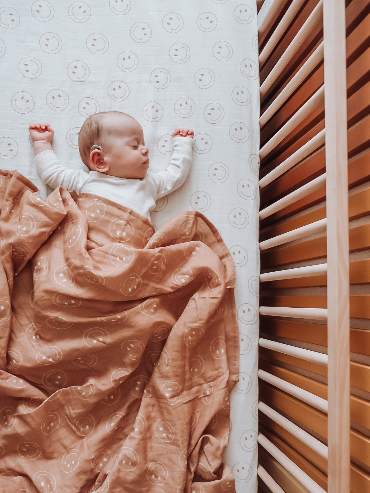 A baby sleeps peacefully on a patterned mattress featuring smiley faces. The baby is cozily enveloped in the Muslin Swaddle | Just Smile Tan by forever french baby, which boasts similar cheerful smiley face designs. Dressed in a white long-sleeve onesie, one arm stretches upwards while the baby's head rests on the mattress beside wooden slats—a scene seemingly lifted from Forever French Baby's dreamy catalog.