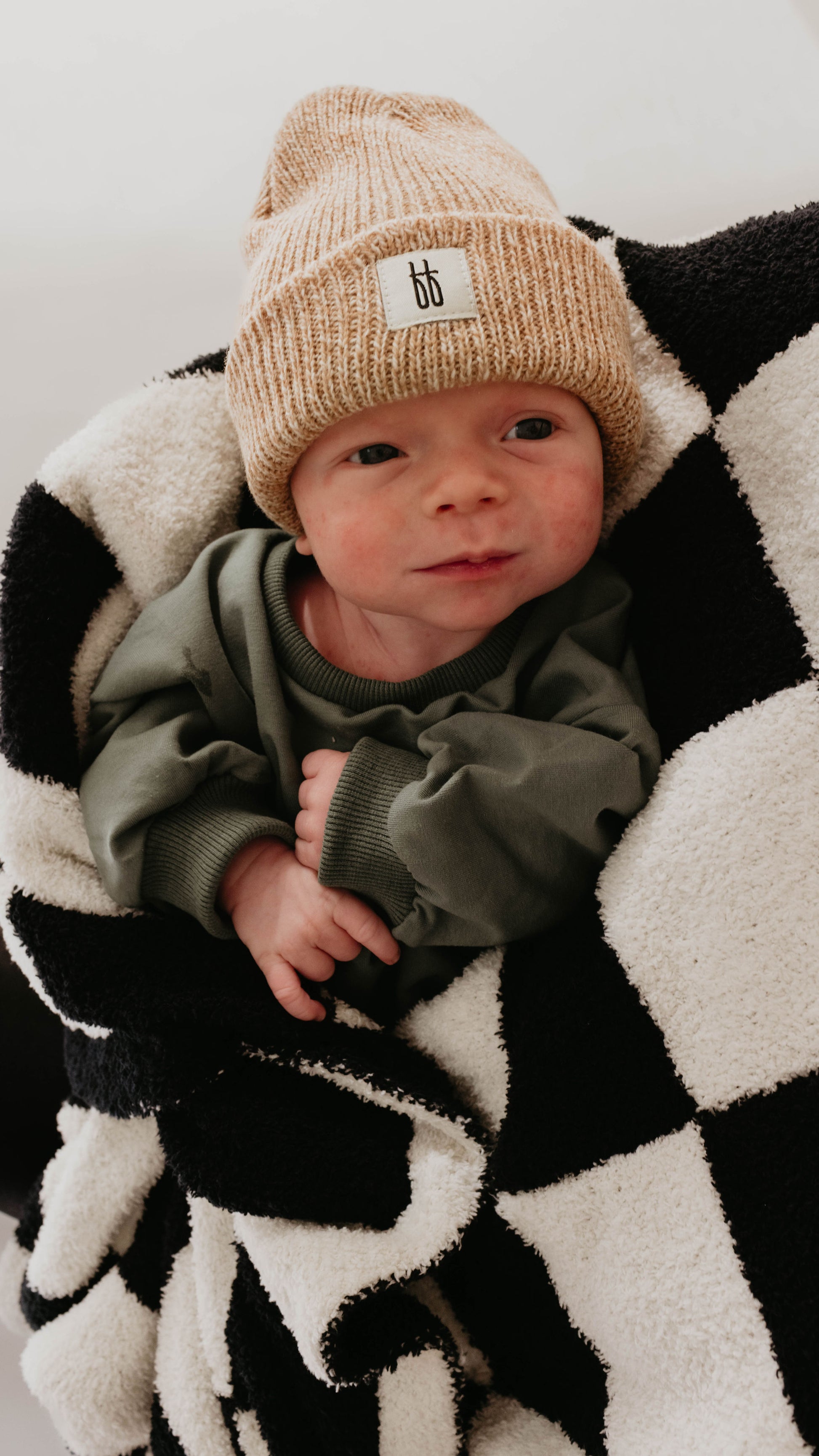 A baby wearing a beige knitted hat and green outfit is wrapped in a soft **Plush Blanket | Black & White Checkerboard** that meets the OEKO Standard 100. The baby looks slightly to the side, with a small smile on their face. The neutral background keeps the focus on this adorable **forever french baby** moment.