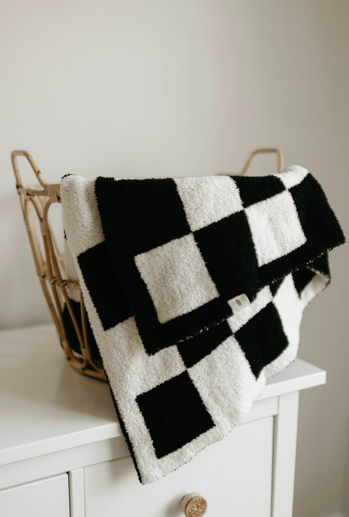 A woven basket sits on top of a white dresser, partially draped with a Forever French Baby Plush Blanket in Black & White Checkerboard. The blanket's bold geometric pattern contrasts with the neutral background, while the basket's woven texture adds a natural element to the scene.