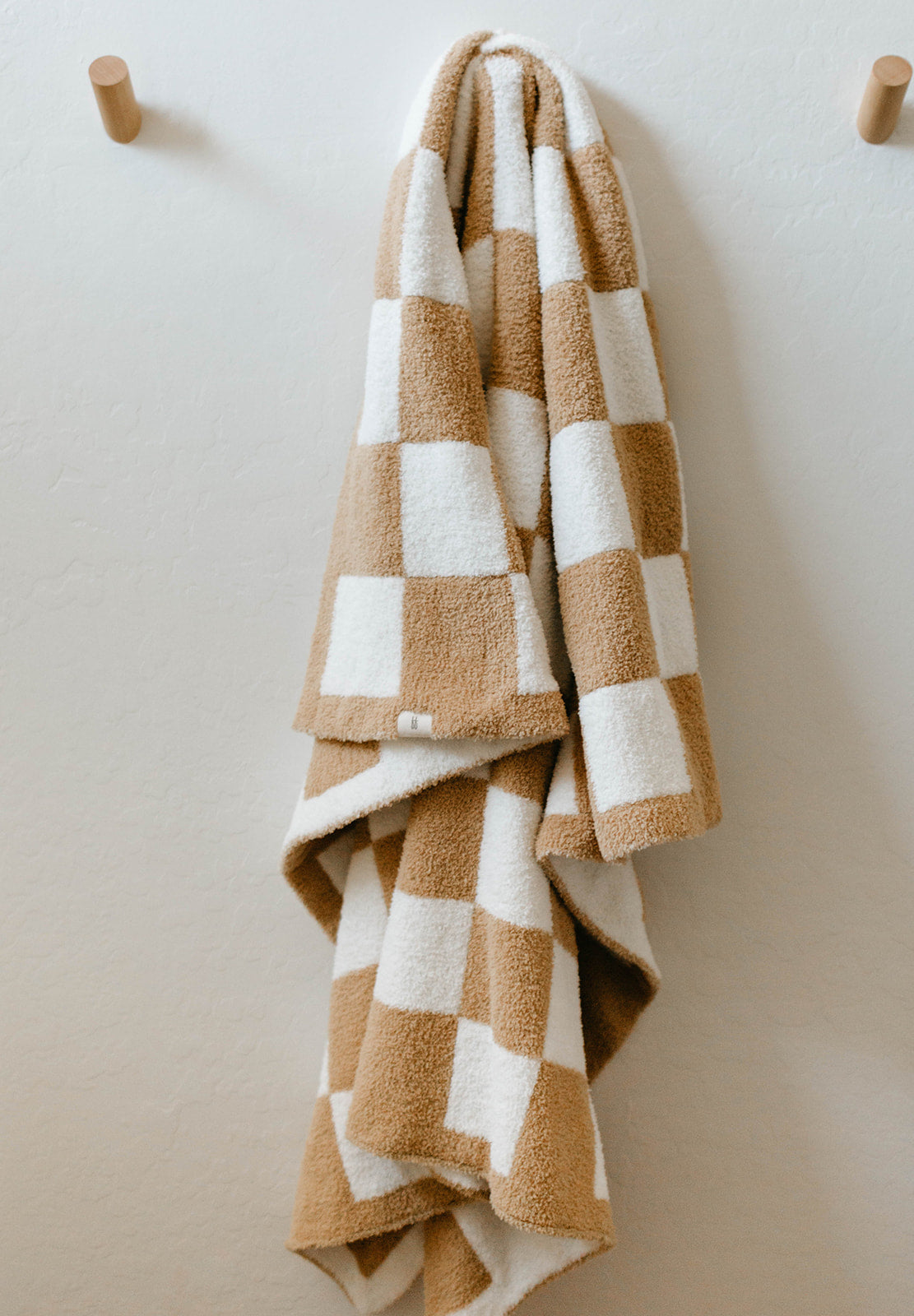 A cozy Plush Blanket | Original Checkerboard by forever french baby, made of microfiber feather yarn with a checkerboard pattern of white and tan squares, hangs on a wooden hook against a light-colored wall. This soft and fluffy blanket is certified by OEKO Standard 100, ensuring top-notch quality and safety.