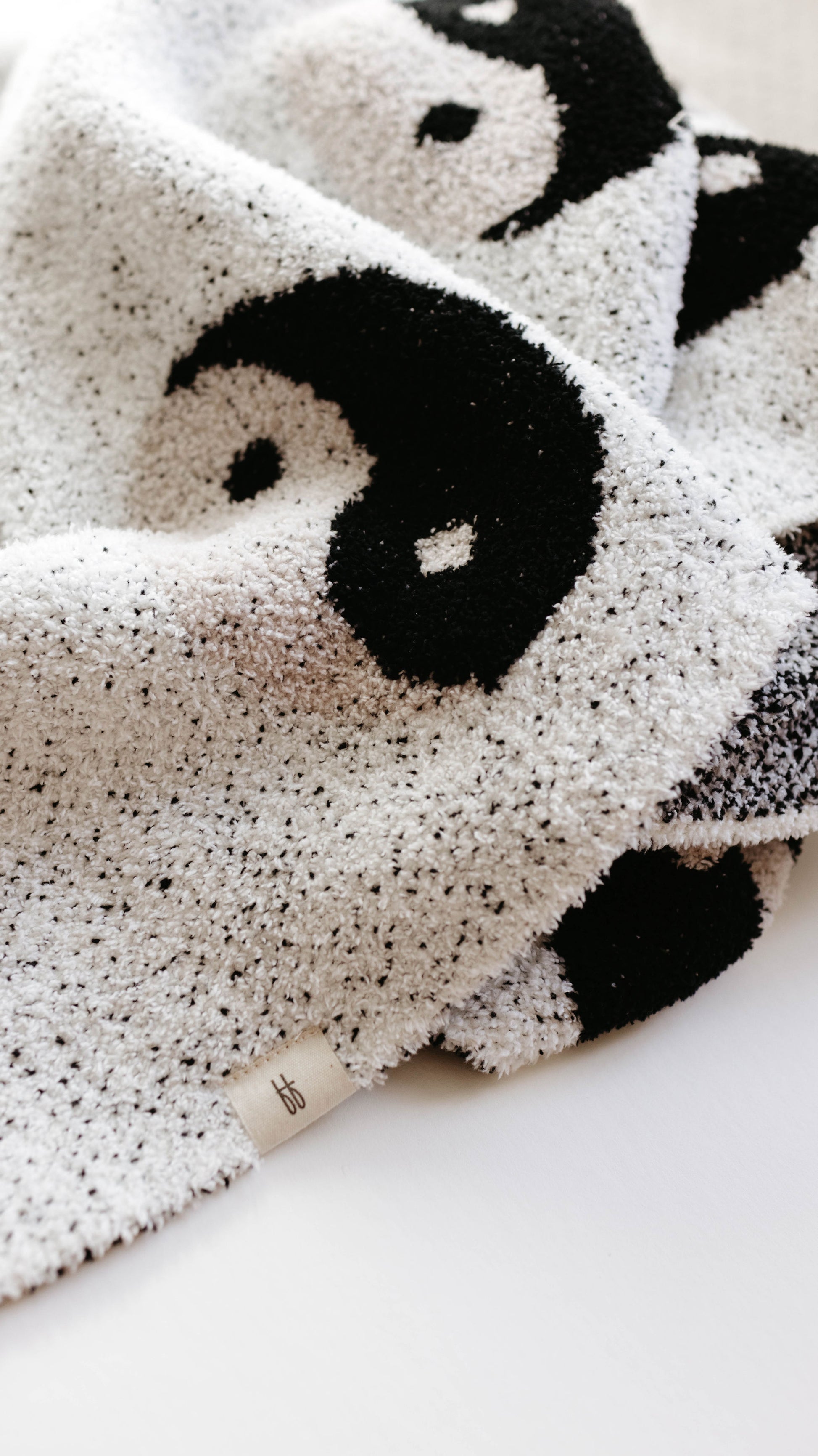 A close-up of the Plush Blanket | Black & White Yin Yang by forever french baby, showcasing its velvety-rich texture and soft, cozy feel. The blanket's striking black and white yin-yang pattern stands out, with a small tag featuring the brand logo near the bottom edge. Made from microfiber feather yarn, it is draped casually to form gentle folds.