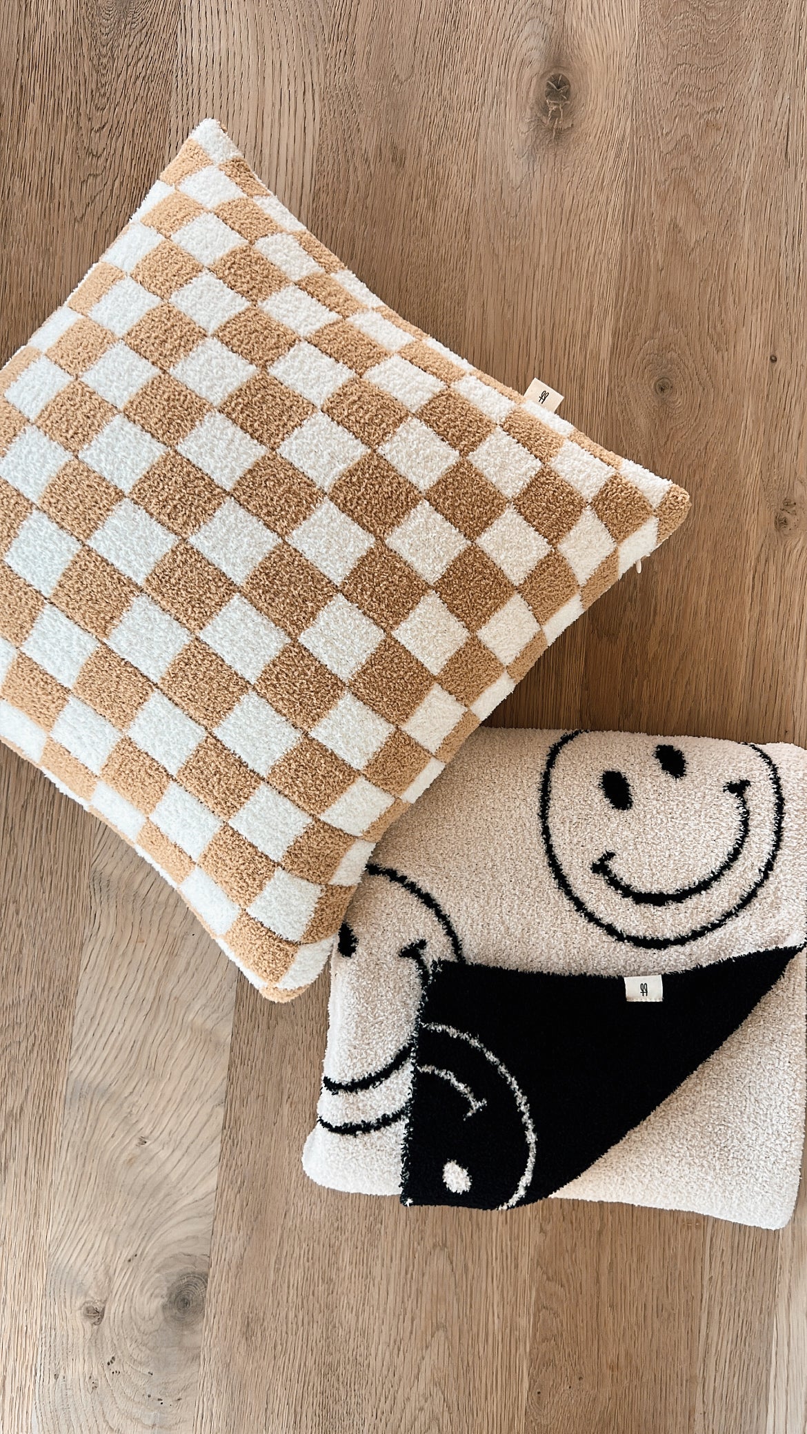 A checkered pillow and a folded blanket lie on a light wooden floor. The pillow, made from Oeko-tex certified materials, has a pattern of alternating beige and white squares. The dreamy soft Plush Blanket | Just Smile Black & White by forever french baby, crafted from microfiber feather yarn, features a smiling face design in black and white. Both items have a cozy, textured fabric.