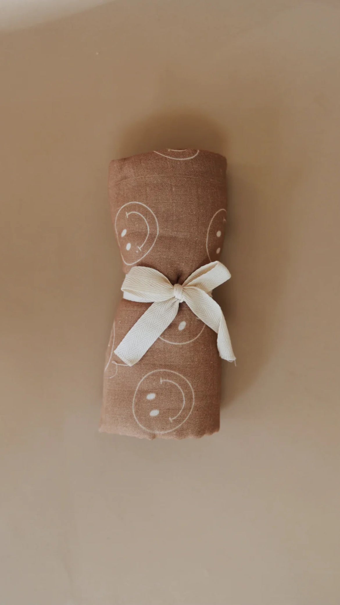 The Just Smile Tan muslin swaddle from forever french baby features a charming brown design adorned with white smiley face patterns and is neatly tied with a white ribbon against a beige background, offering perfect comfort for your little one.