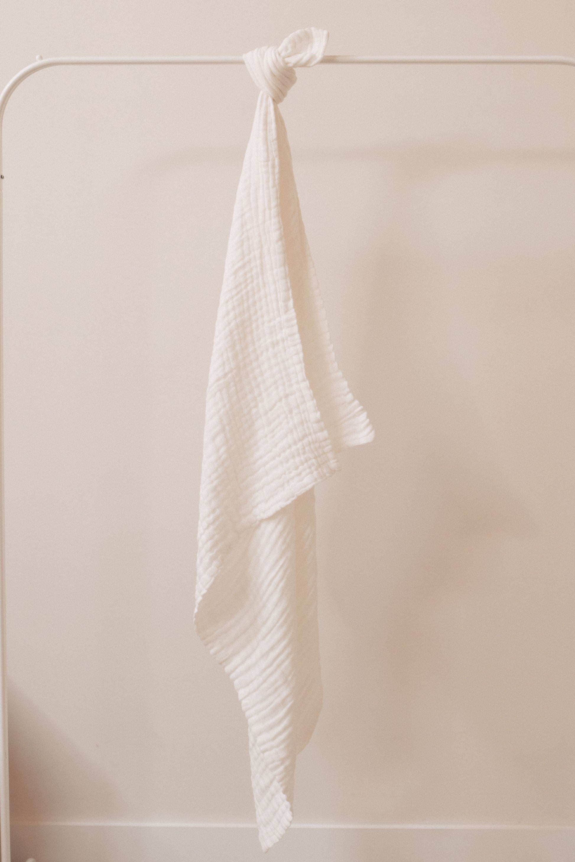 A soft, beige-colored Crib Sheet from forever french baby, named "Cloud," is draped and knotted over a simple white metal clothing rack. The background features a plain, off-white wall, creating a minimalist and serene composition. Remember to wash it with like colors and tumble dry on low to maintain the fabric’s gentle texture.