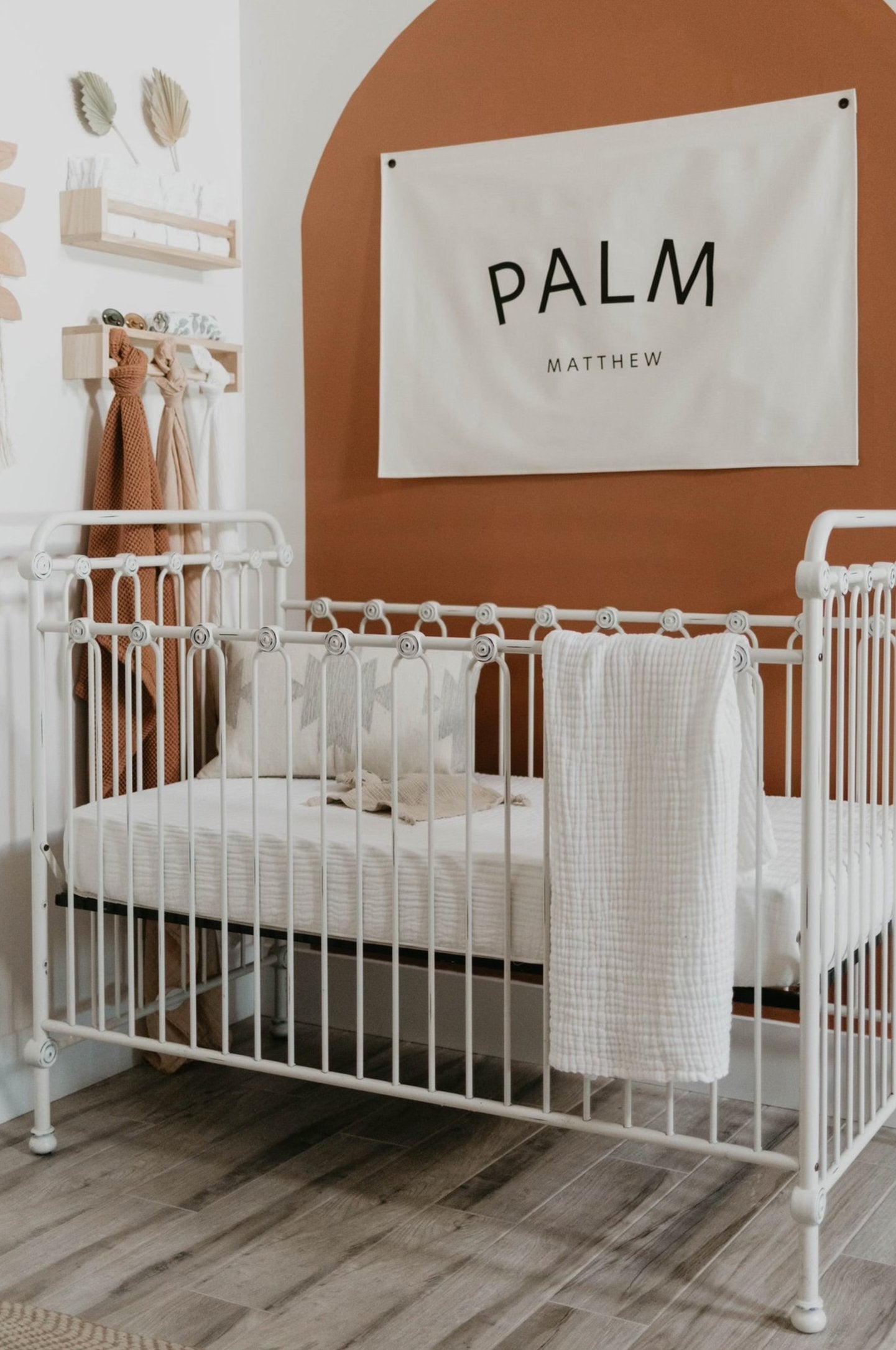 A cozy nursery includes a white metal crib outfitted with white bedding from Forever French Baby's Crib Sheet | Cloud collection, and a blanket draped over the side. Behind the crib, a terracotta-colored arch on the wall displays a banner that reads "Palm Matthew." The room boasts wooden floors and soft decor elements.