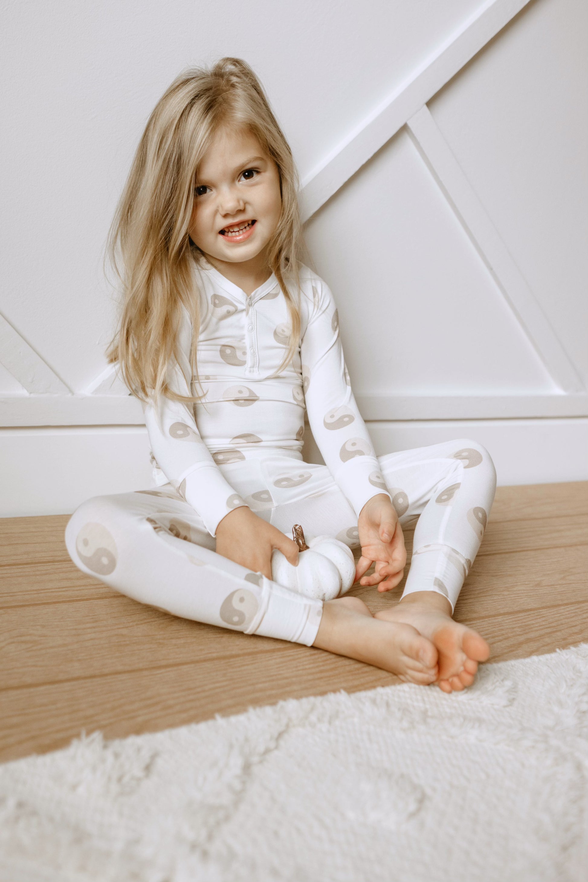 A young child with long blonde hair sits on a wooden floor in a white room. Wearing forever french baby's hypo-allergenic Yin Yang Tan and White Bamboo Two Piece Pajamas featuring a yin-yang pattern, they hold a small white pumpkin. A soft, white rug is partially visible in the foreground.