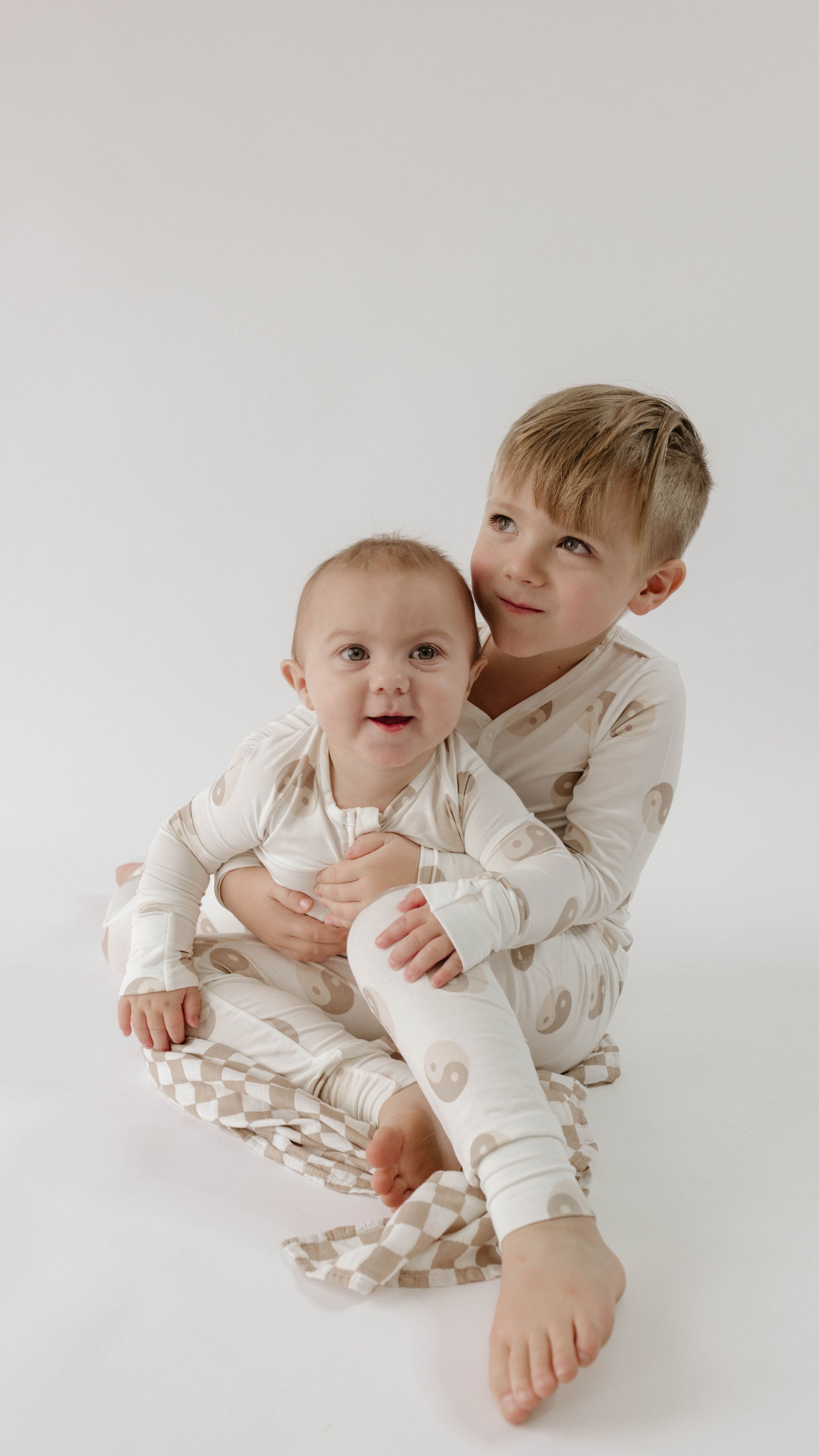 Two young children sit together in matching Forever French Baby "Yin Yang Tan and White" Bamboo Two Piece Pajamas against a plain white background. The older child smiles while sitting behind the younger sibling and hugging them. Both appear content and happy, enjoying their hypo-allergenic clothing.