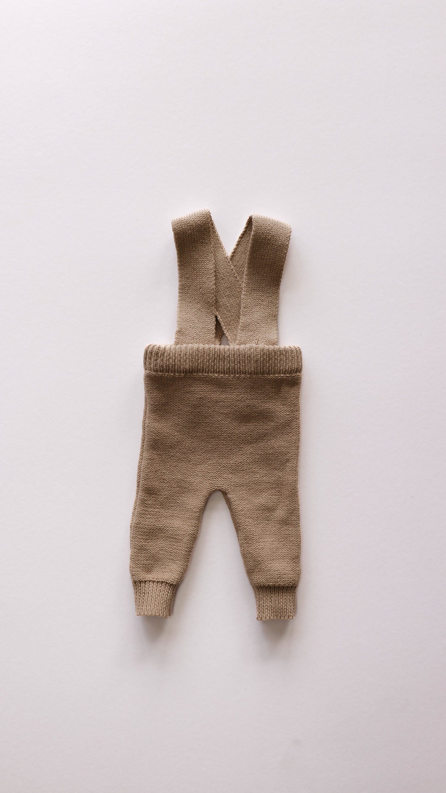 A pair of Forever French's Knit Suspenders | Graham, beige knitted baby pants, lies flatly on a light gray background. Perfect for cooler weather, the suspenders are crossed at the back and the fabric features a ribbed texture around the waist and ankles.