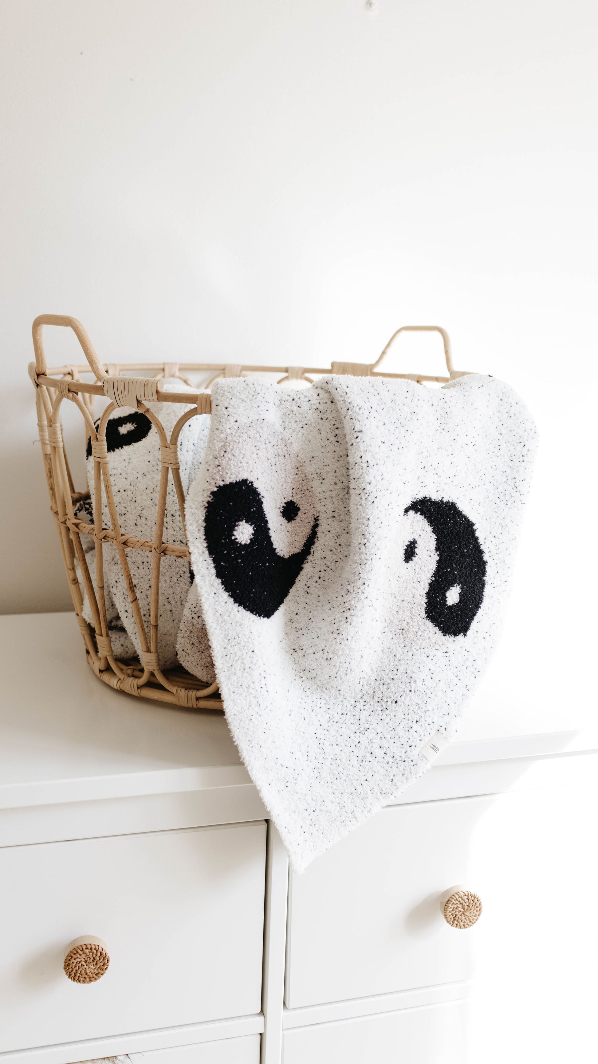 A wicker basket sits on top of a white dresser. Two white Plush Blankets by forever french baby, made from velvety-rich microfiber feather yarn and adorned with black yin-yang symbols, are draped over the edge of the basket, with one blanket hanging further down. The background is a plain white wall.