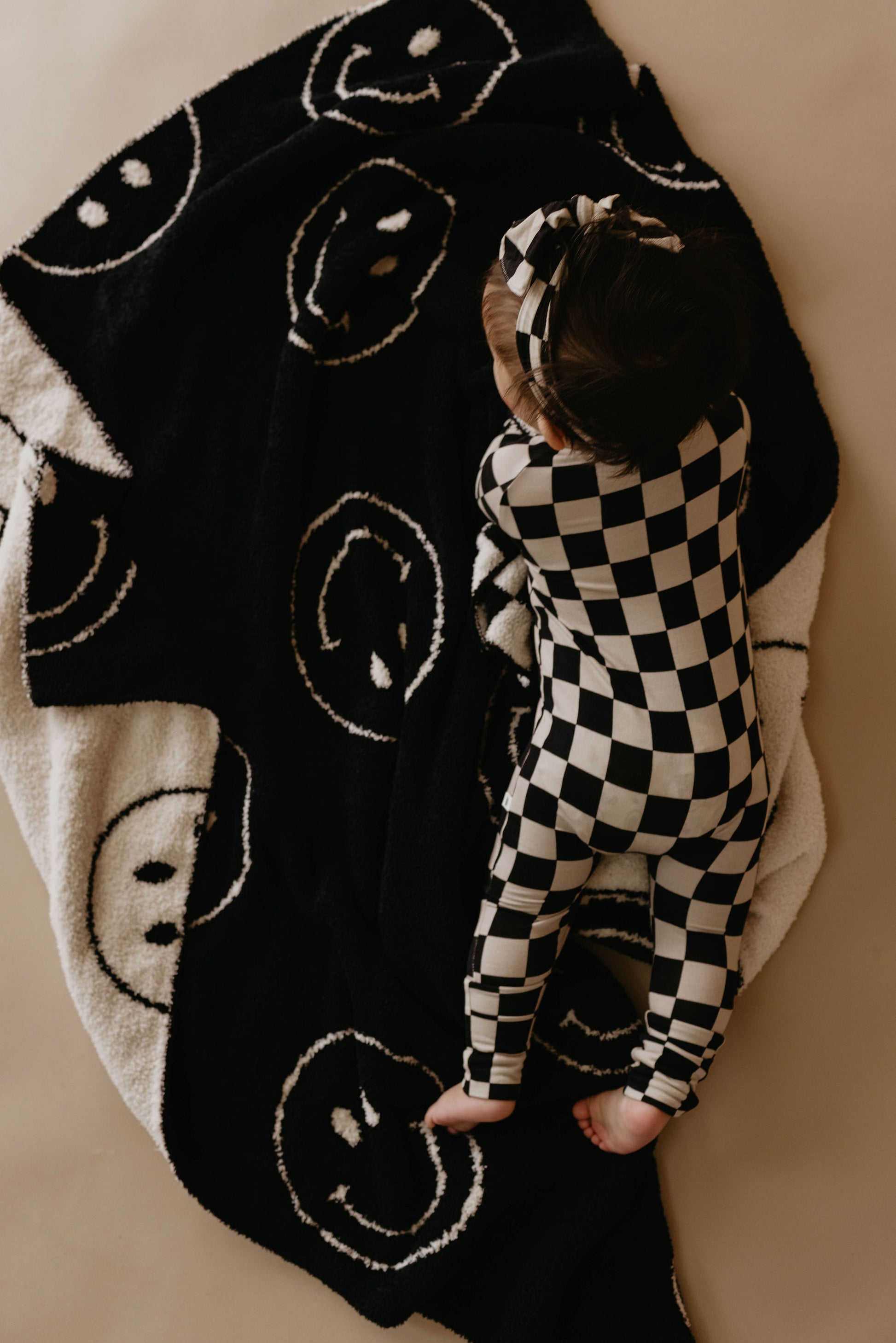 A baby wearing a black-and-white checkered outfit and matching headband lays on their stomach on the Plush Blanket | Just Smile Black & White by forever french baby. The photo, taken from above, captures the baby peacefully resting on the dreamy soft blanket made of Microfiber feather yarn with smiley face patterns.