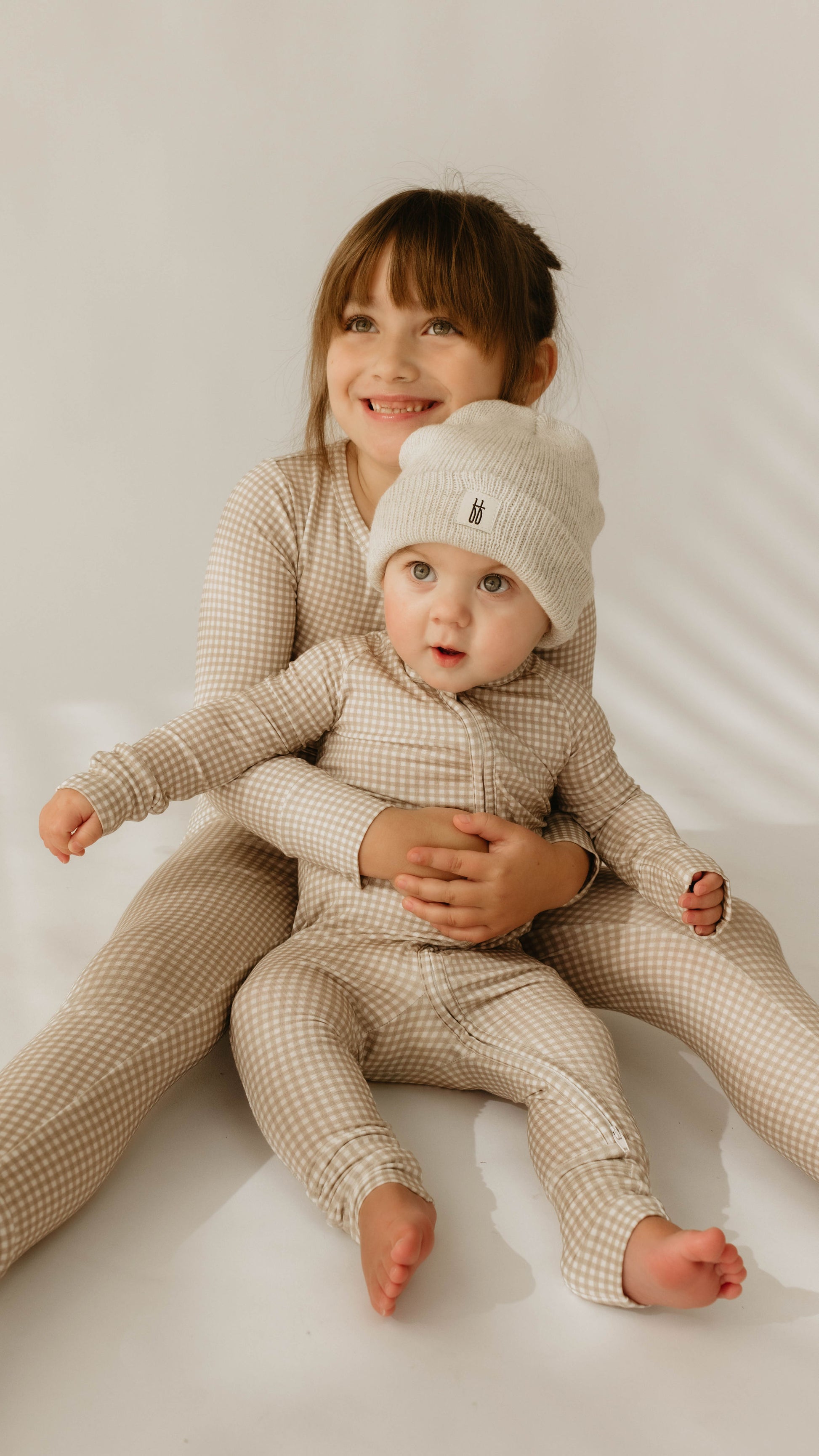 Two young children are sitting on the floor, both dressed in matching Ryan Gingham Bamboo Two Piece Pajamas from forever french baby. The older child is smiling and holding the baby, who is also wearing a beanie. Both are looking towards the camera. The background is minimal and softly lit.